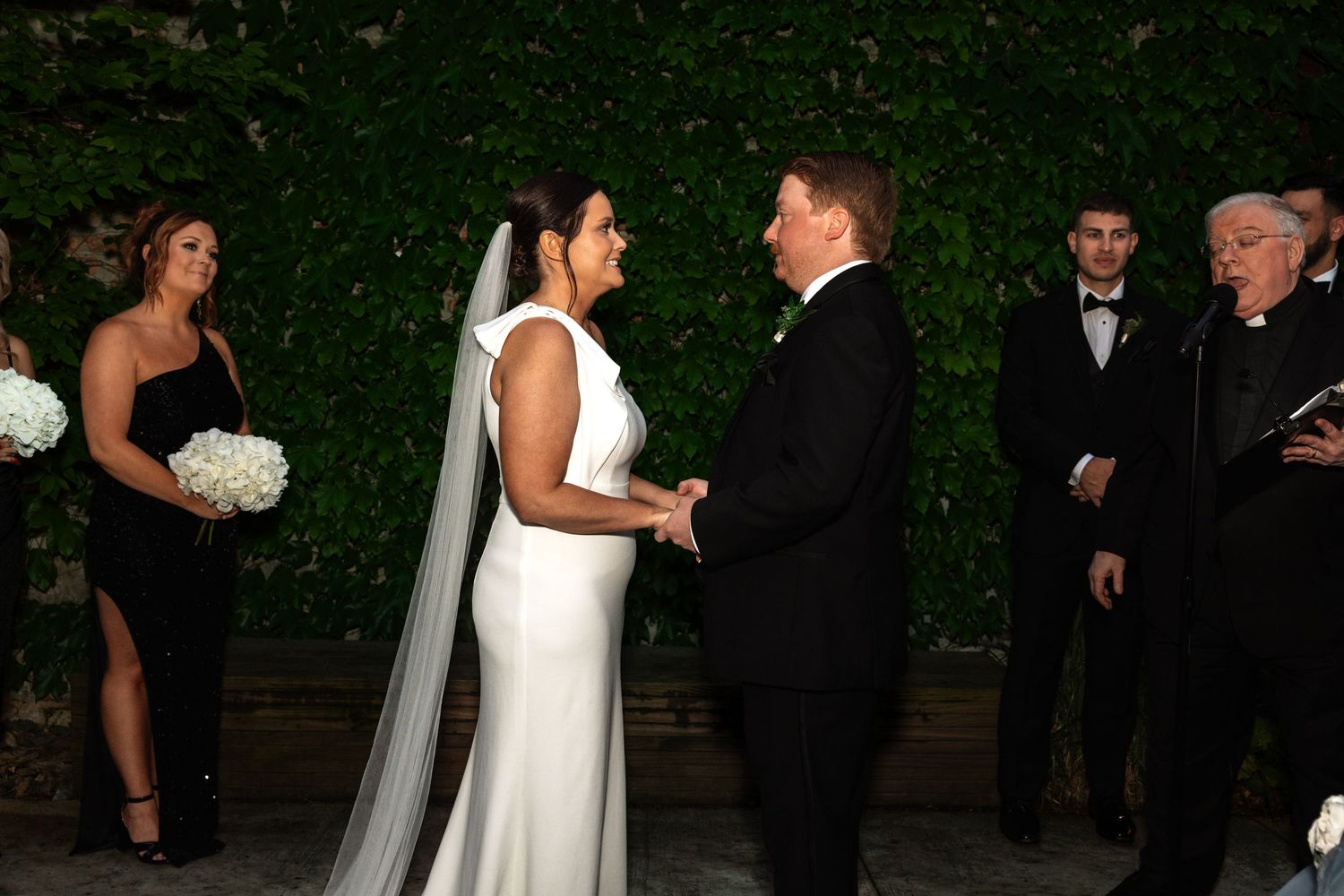 Smiling bride and groom hold hands at outdoor wedding ceremony with bridal party.