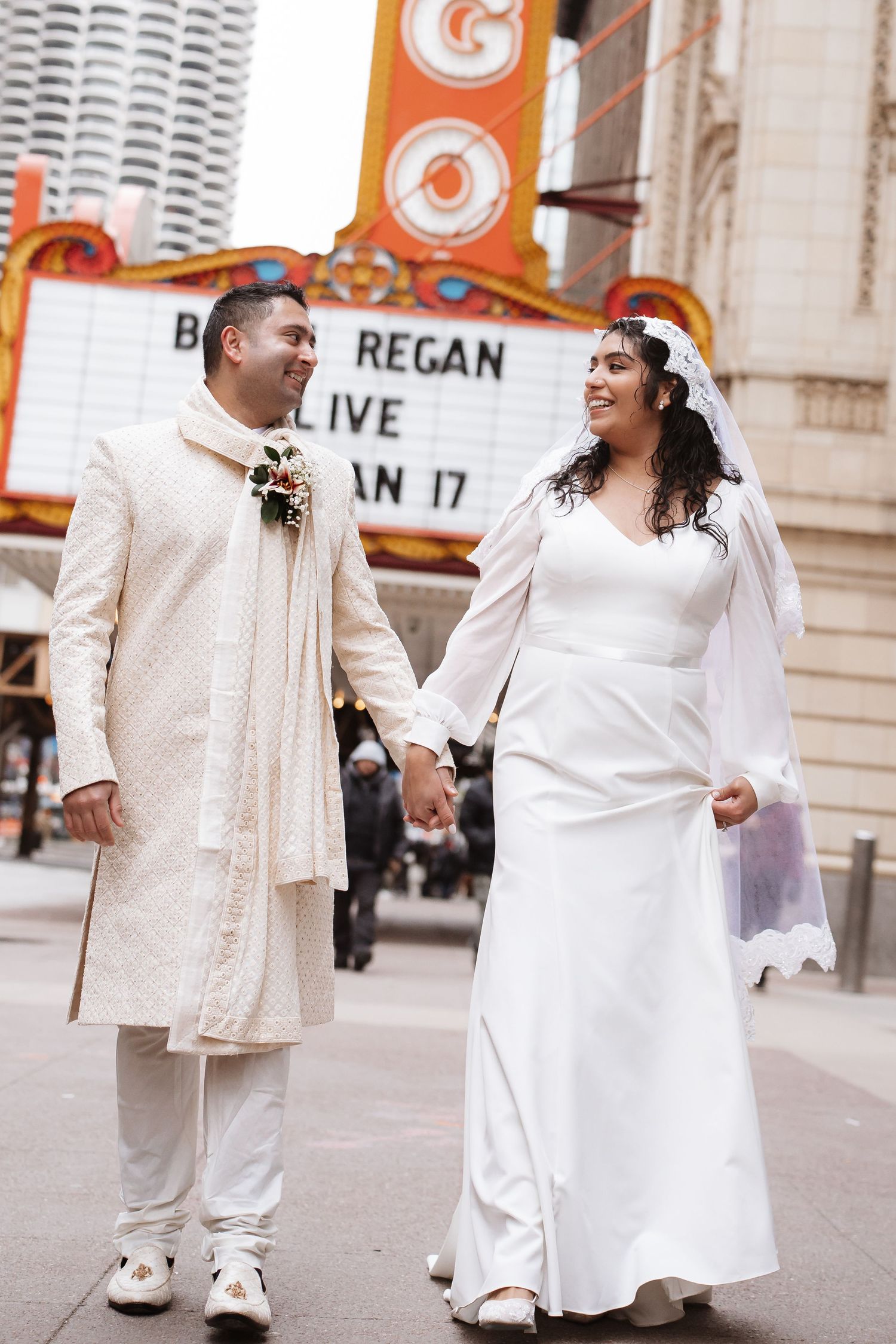 Smiling South Asian newlyweds walk hand-in-hand past Chicago Theatre Brian Regan marquee.