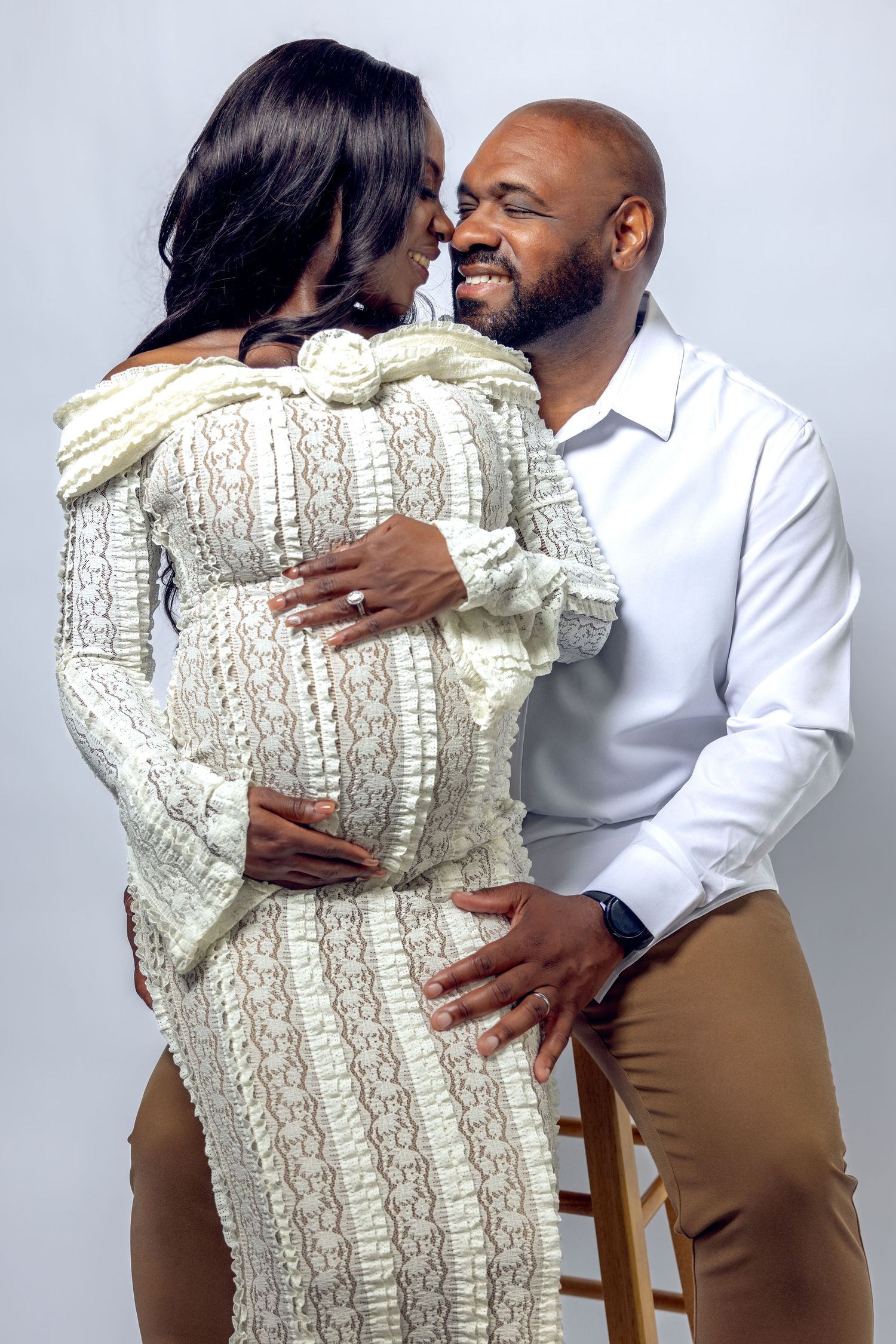 Expecting couple sharing a close moment from behind, man on stool embracing woman in white lace dress.