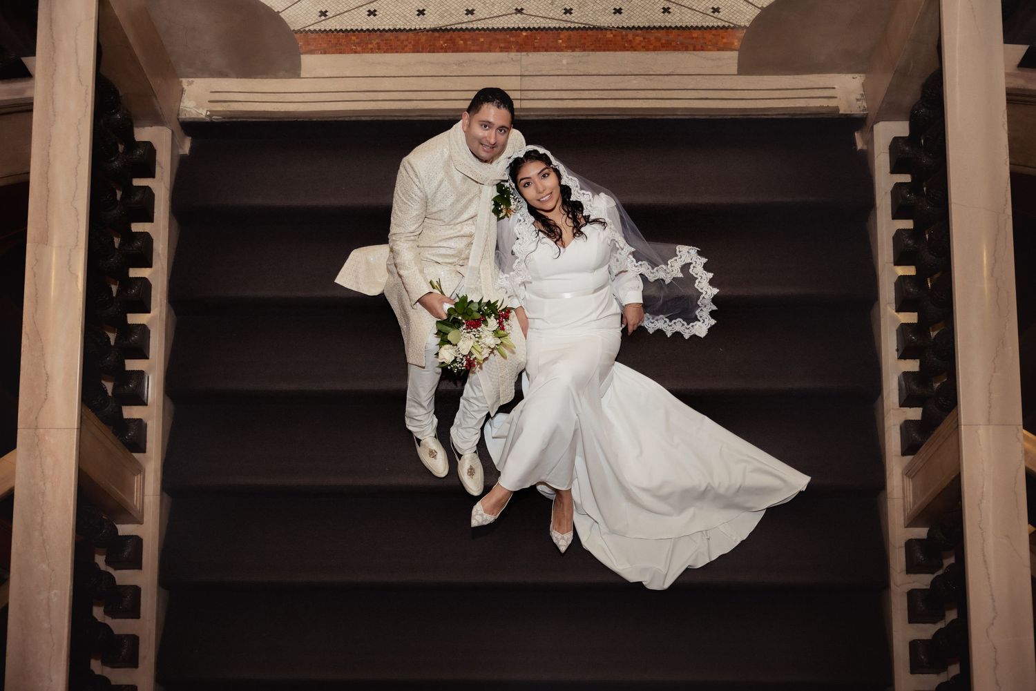 Aerial view of bride and groom sitting on dark staircase, bride holding floral bouquet.