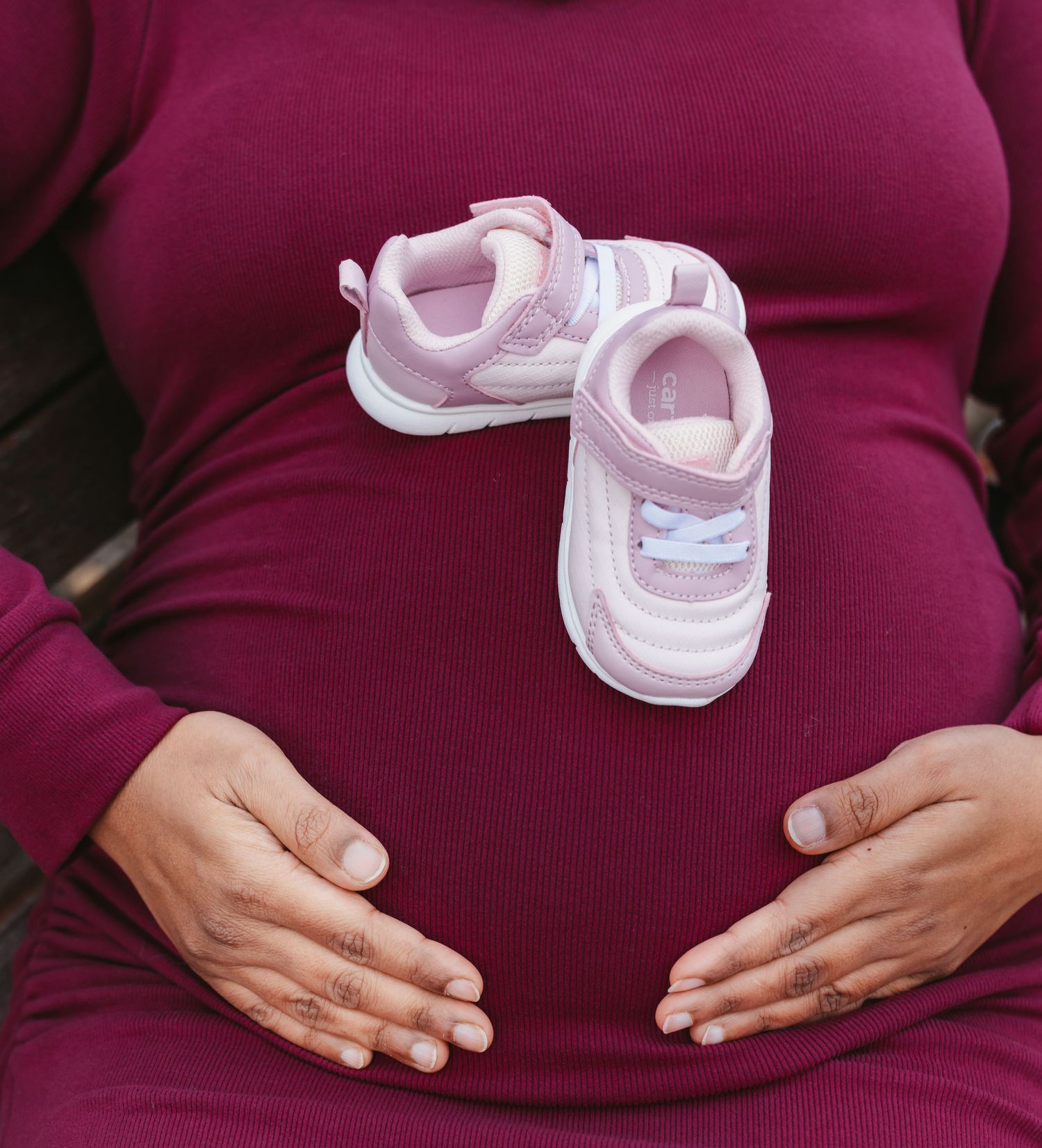 Close-up of pregnant woman in burgundy dress holding pink baby sneakers on her baby bump.