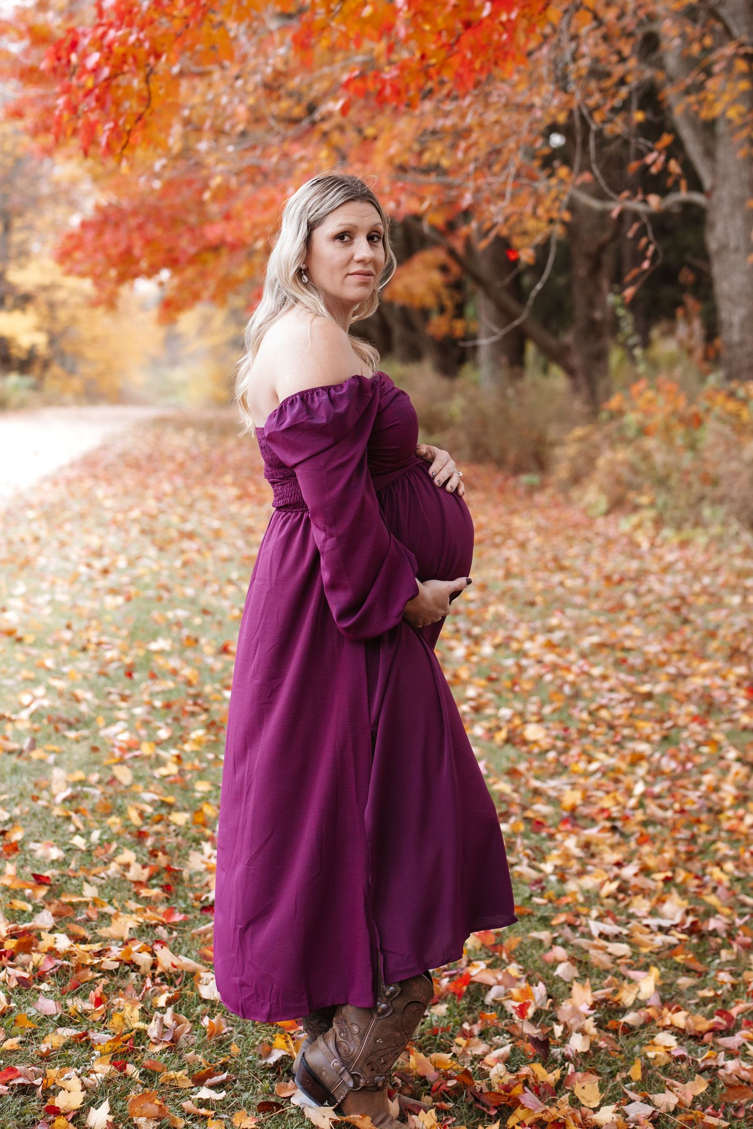 Pregnant woman in purple off-shoulder dress poses outdoors among vibrant autumn foliage.