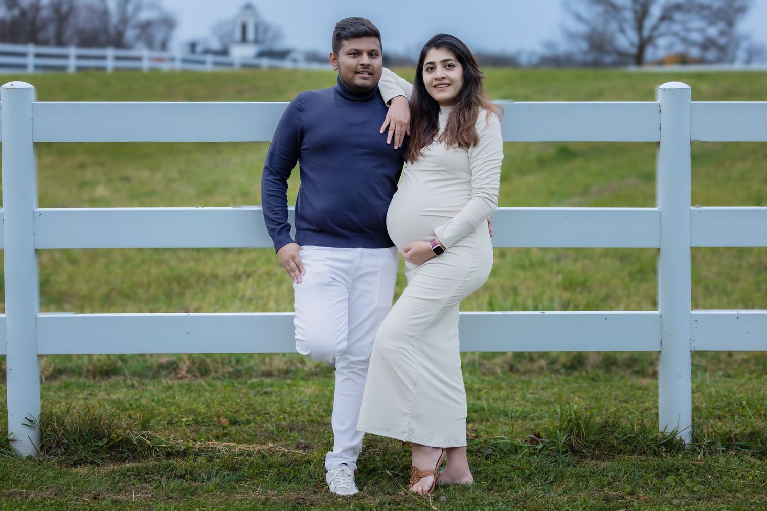 Expecting couple in white and navy outfits posing by a white fence on a farm.