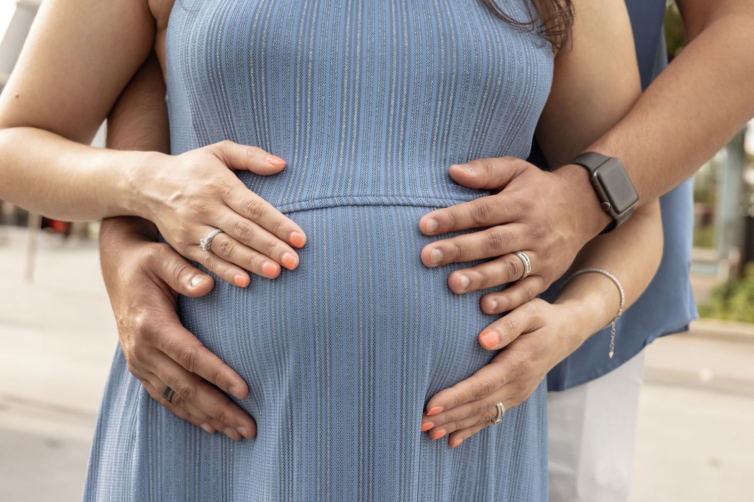 Close-up of two pairs of hands cradling a pregnant belly in a light blue maternity dress outdoors.