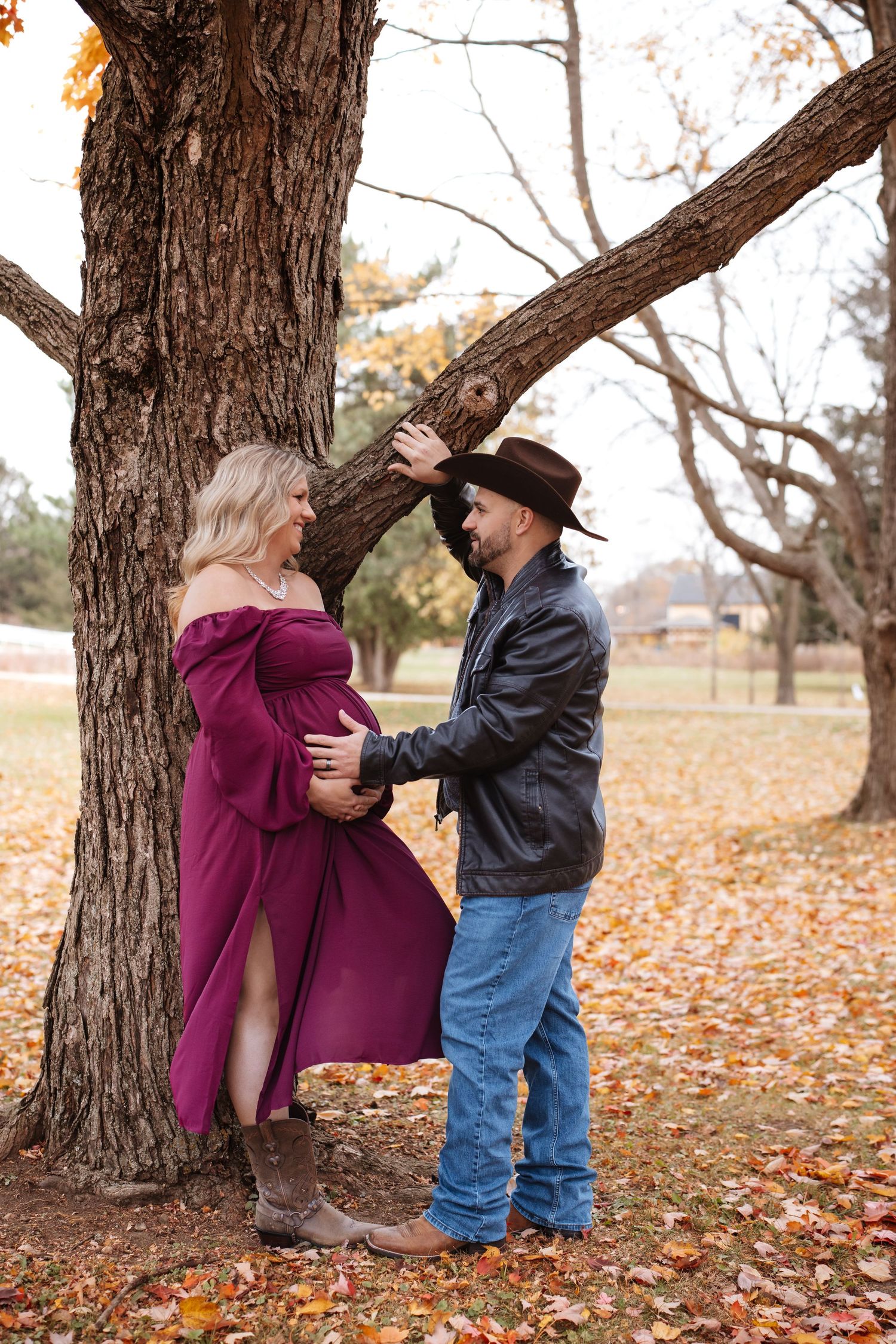 Pregnant woman in burgundy gown with cowboy partner touching her belly by autumn tree.