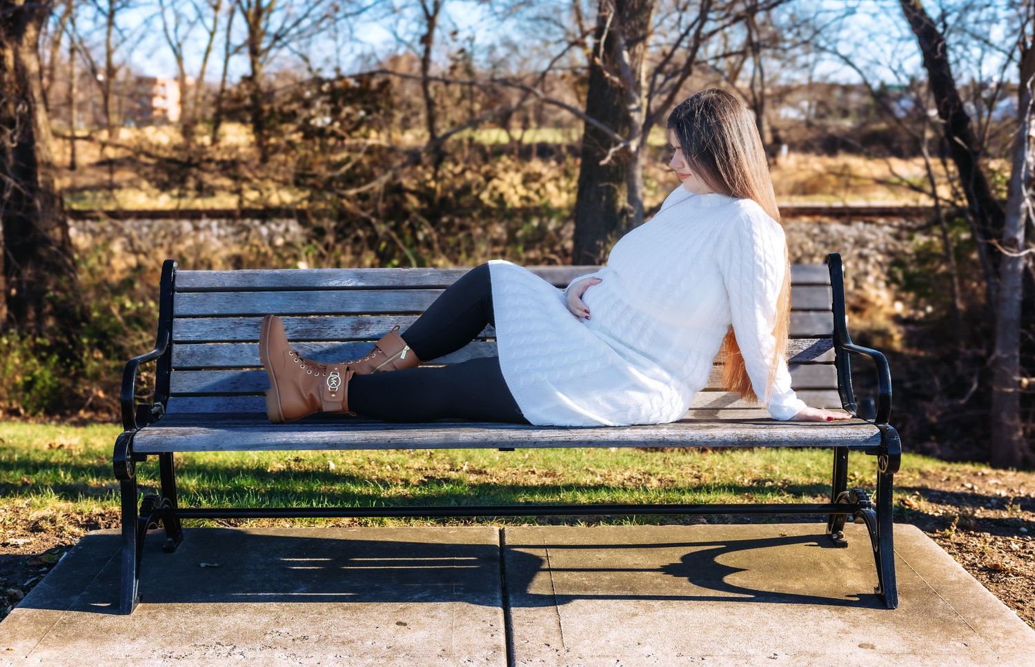 Pregnant woman in white sweater lying on park bench, relaxing outdoors in autumn setting.