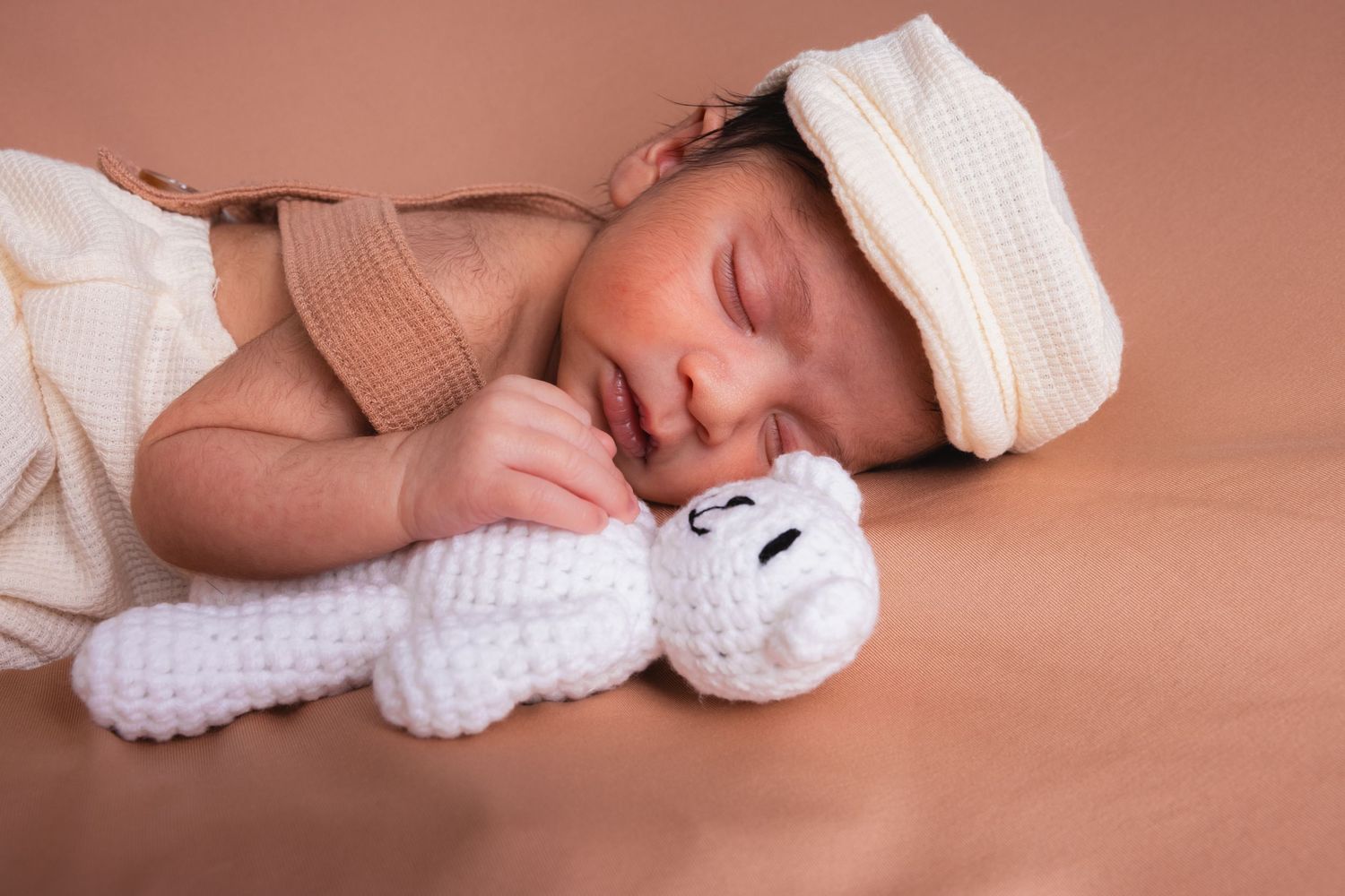 Close-up of sleeping newborn baby with white knitted teddy bear on warm beige background.