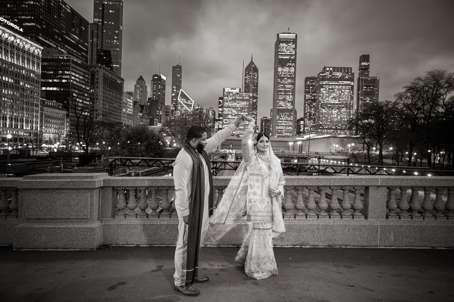 Black and white photo of couple in traditional South Asian wedding attire with Chicago skyline at night.