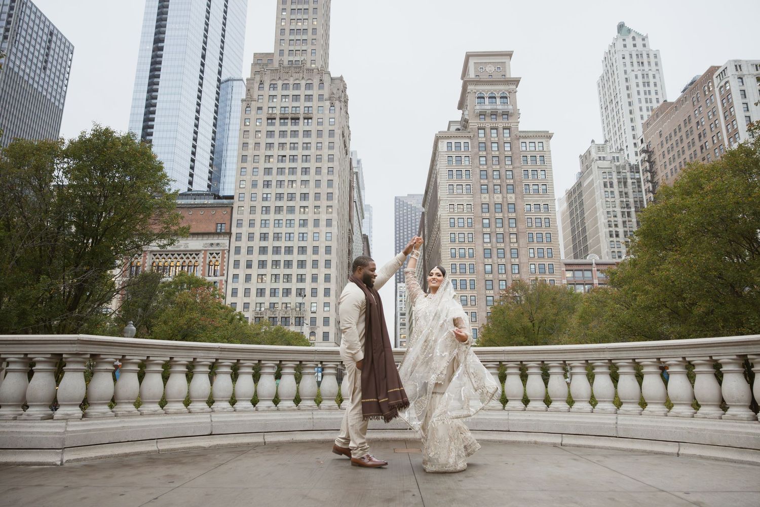 Couple in traditional South Asian wedding clothes dance on bridge with Chicago skyline at dusk.