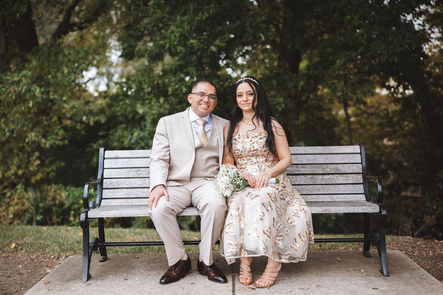 Smiling newlywed couple sitting together on park bench in elegant wedding attire.