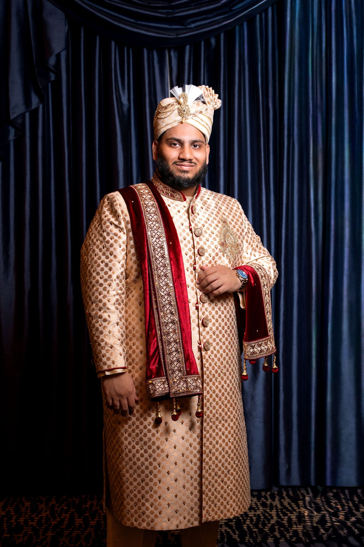 Groom in ornate gold sherwani and turban posing confidently before blue velvet curtain backdrop.