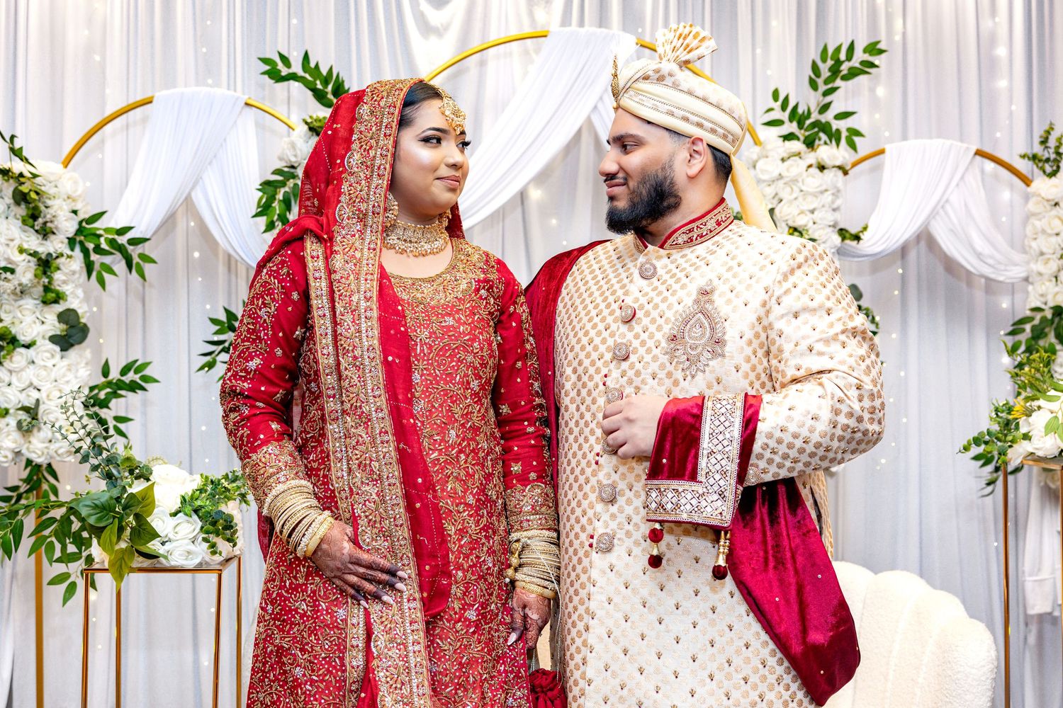 South Asian wedding couple in red and gold traditional attire sharing a loving glance at ceremony.