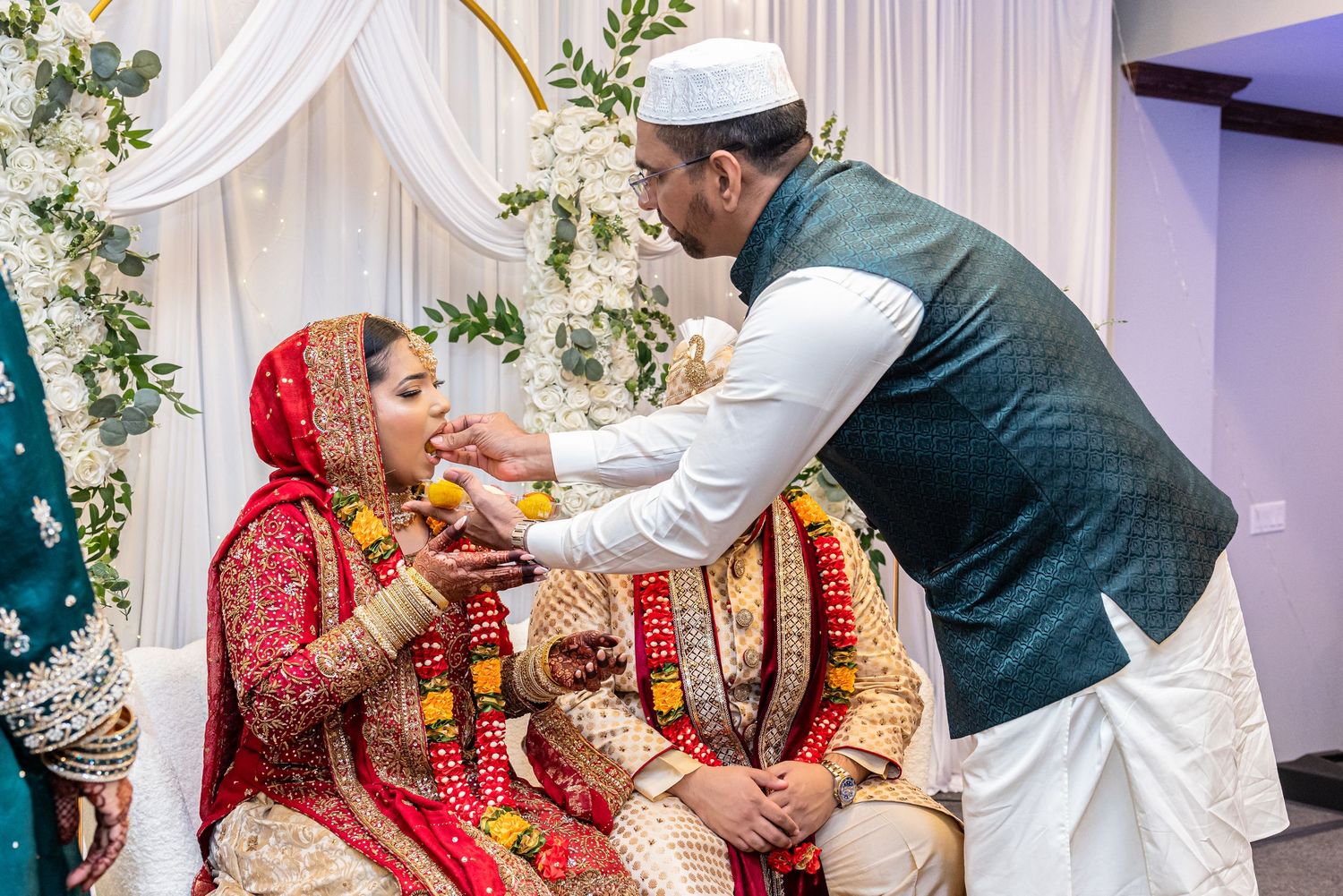 Man in white kufi feeds bride in red bridal attire during South Asian wedding ceremony.