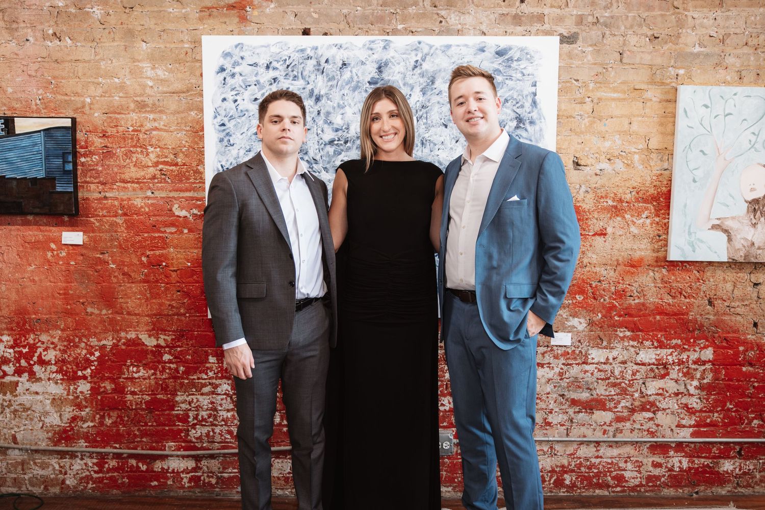 Three smiling professionals in formal attire pose at an upscale art gallery opening event.