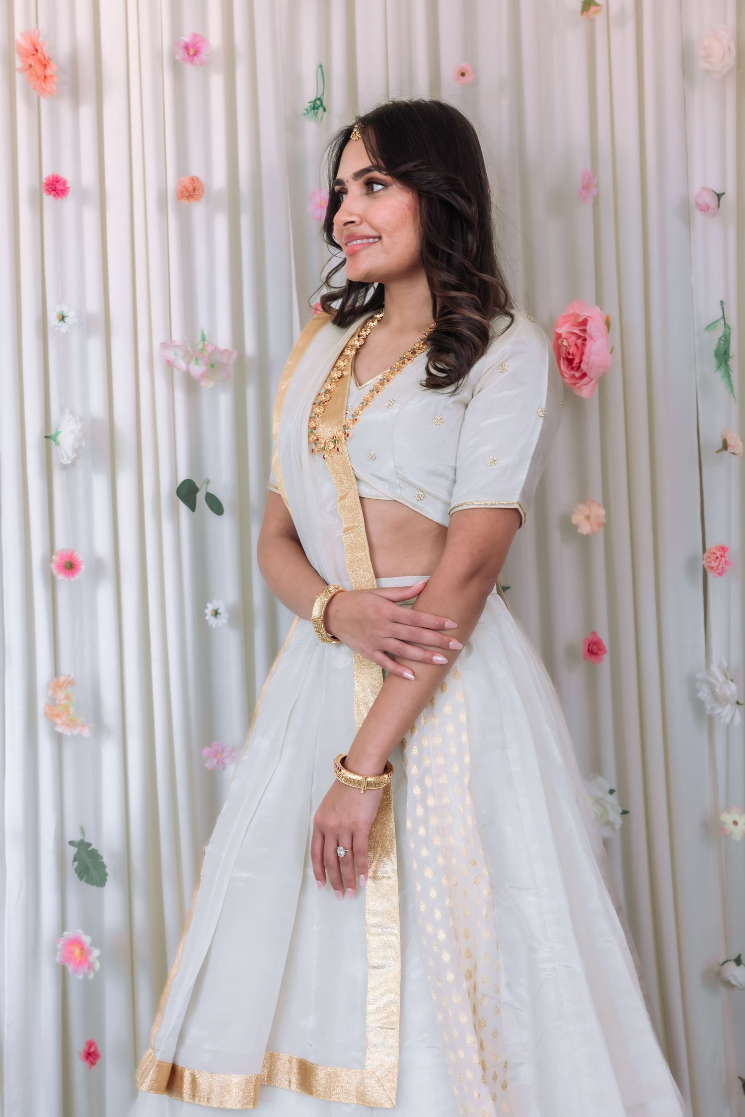 Beautiful woman in white lehenga with gold border smiling against a floral decorated backdrop.