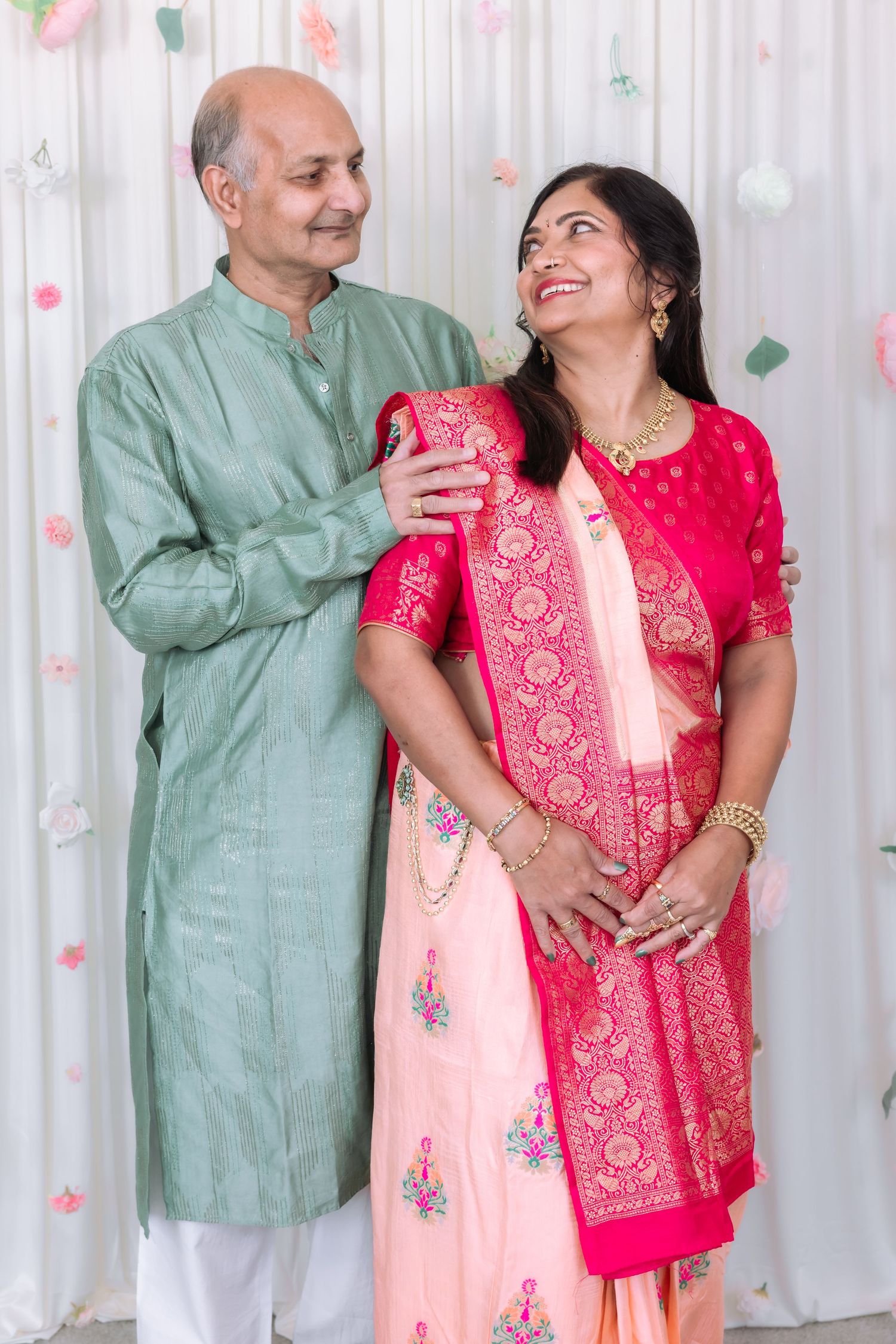 Indian couple in traditional attire share a joyful moment together at a floral decorated white wedding backdrop.
