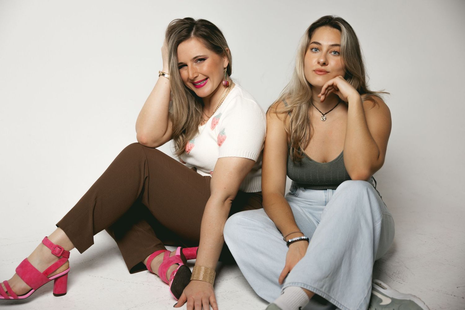 Two blonde women sitting together on white floor in casual outfits posing for studio photo.