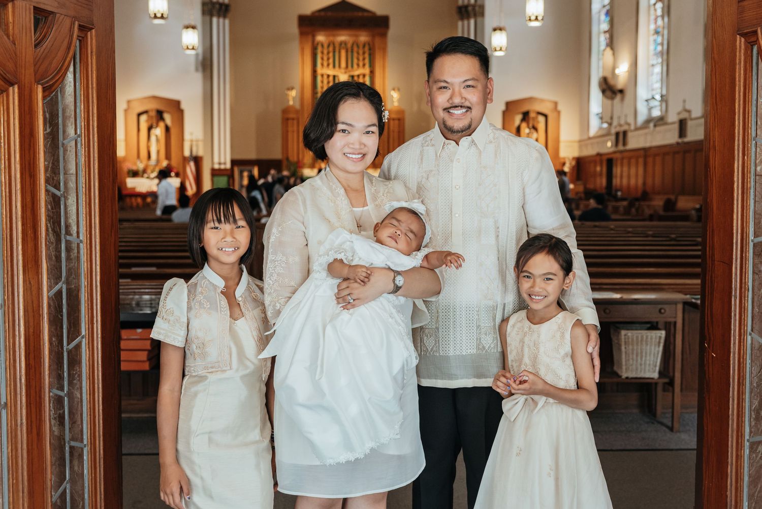 Smiling Filipino family in white outfits poses inside church during baby baptism celebration.