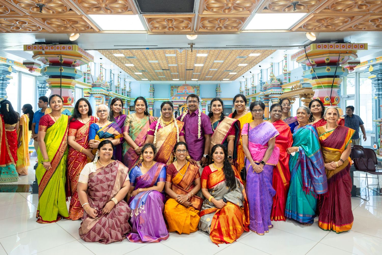 Large group of women in colorful South Indian silk sarees posing together at a Hindu temple.