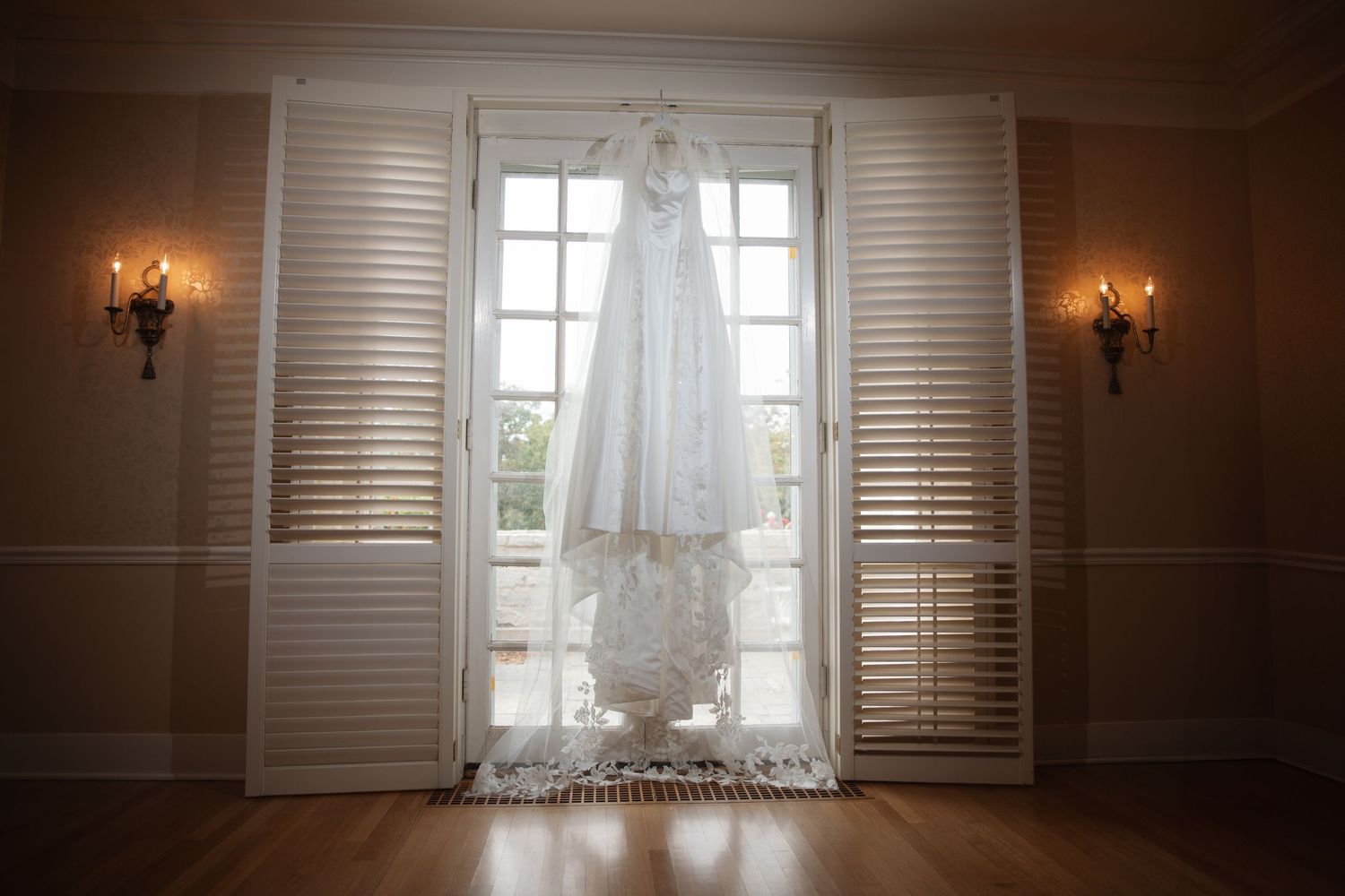 White lace wedding dress hanging in a doorway framed by shuttered panels and decorative wall sconces in dim lighting.