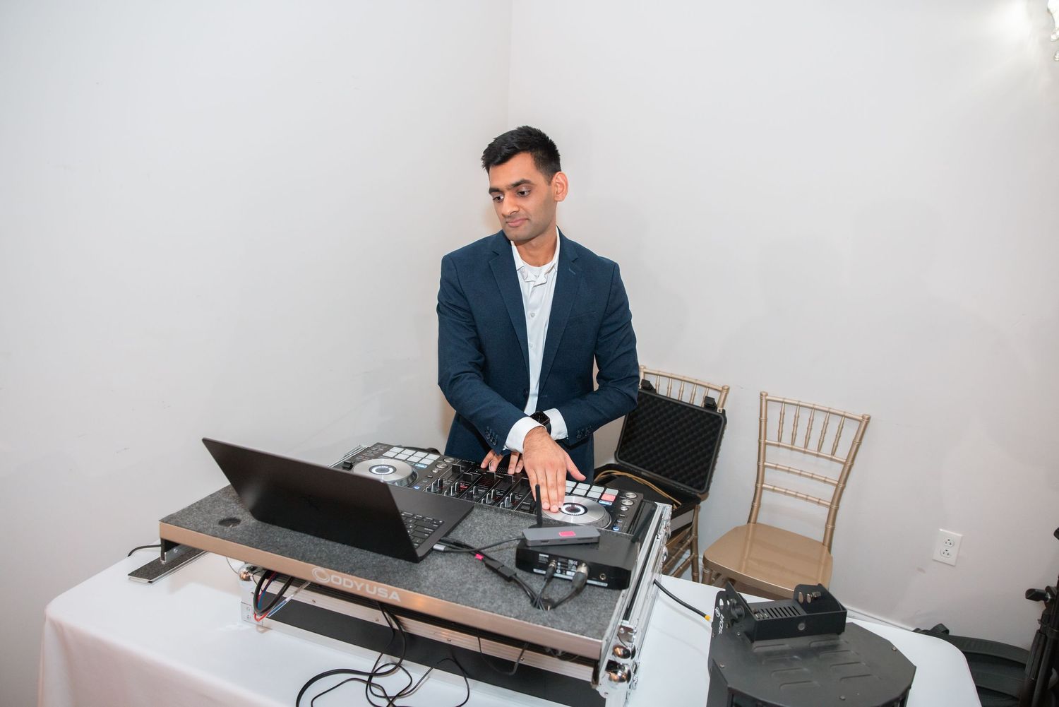DJ in a navy suit operating mixing equipment and laptop at a white draped table during an event.