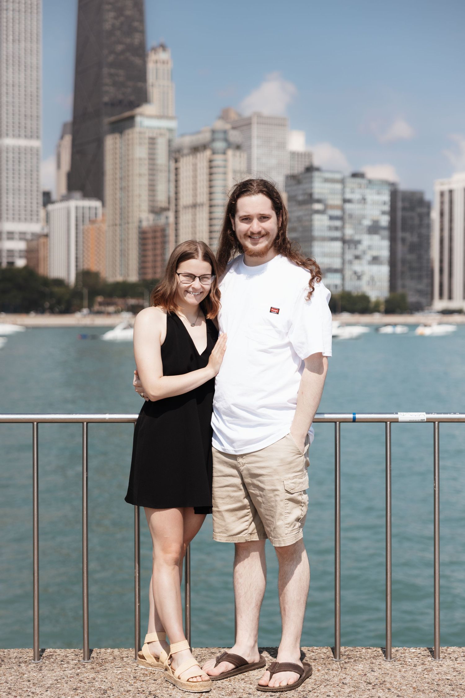 Young couple in casual summer outfits posing by Lake Michigan with Chicago skyline behind them.