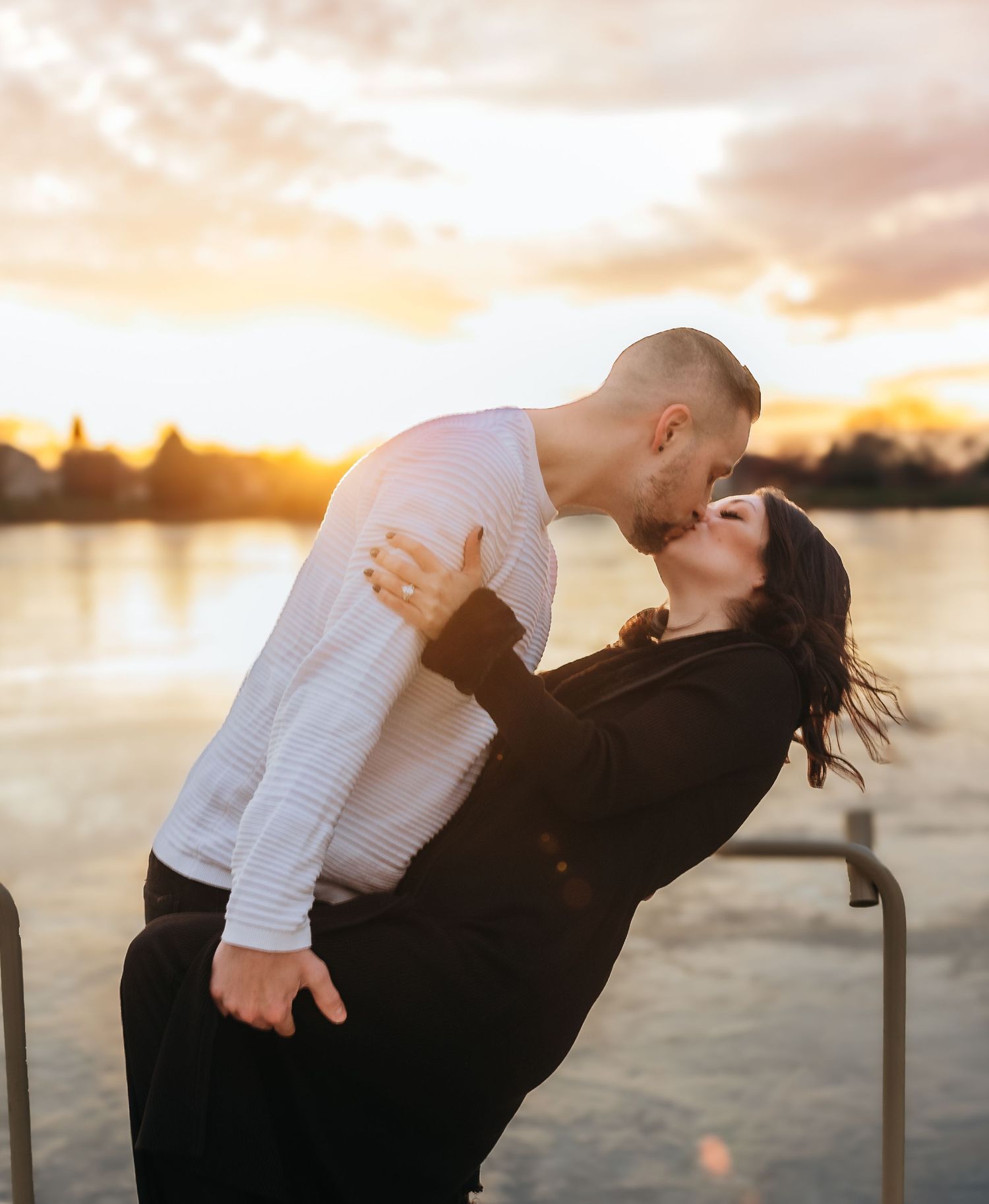 Couple sharing a passionate dip kiss at sunset by a waterfront, golden light illuminating the romantic scene.