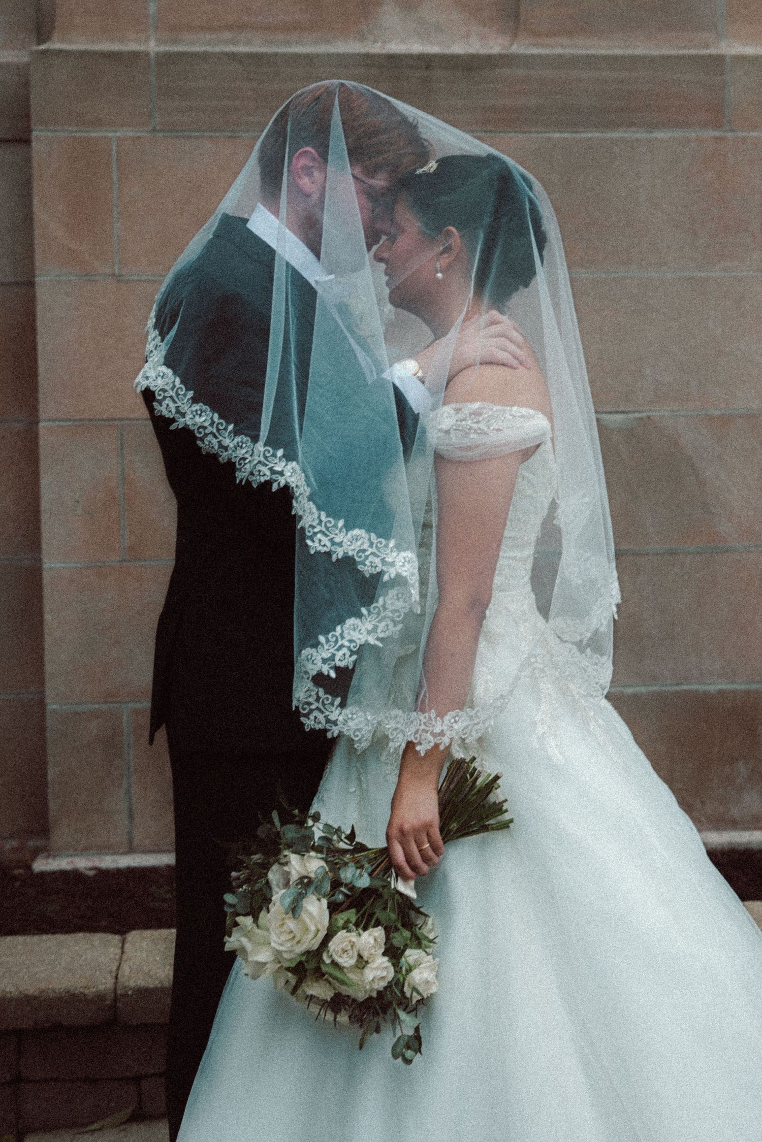 Bride and groom touching foreheads under veil, bride holding white bouquet.