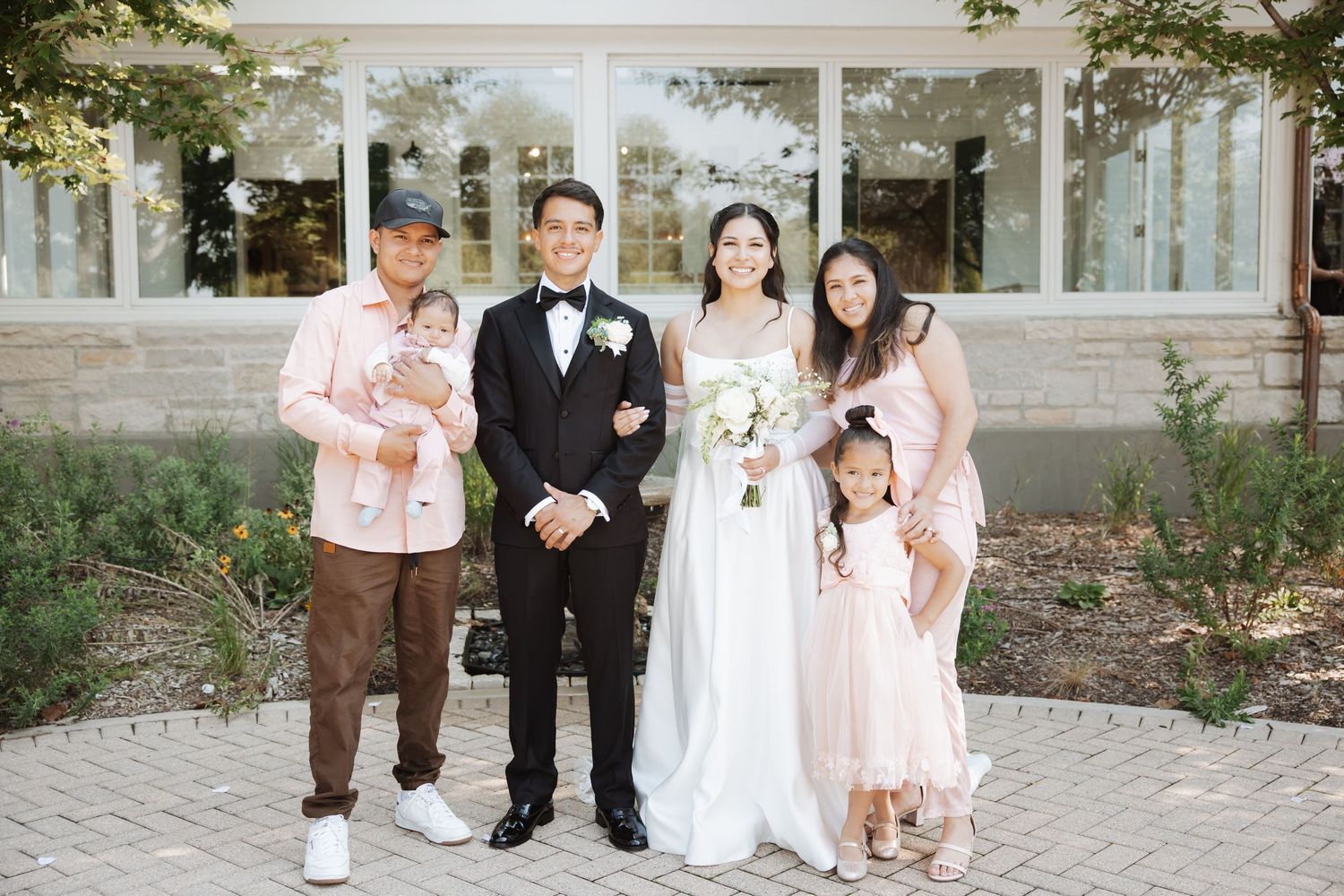 Bride and groom pose with small family including two young children outside modern wedding venue.