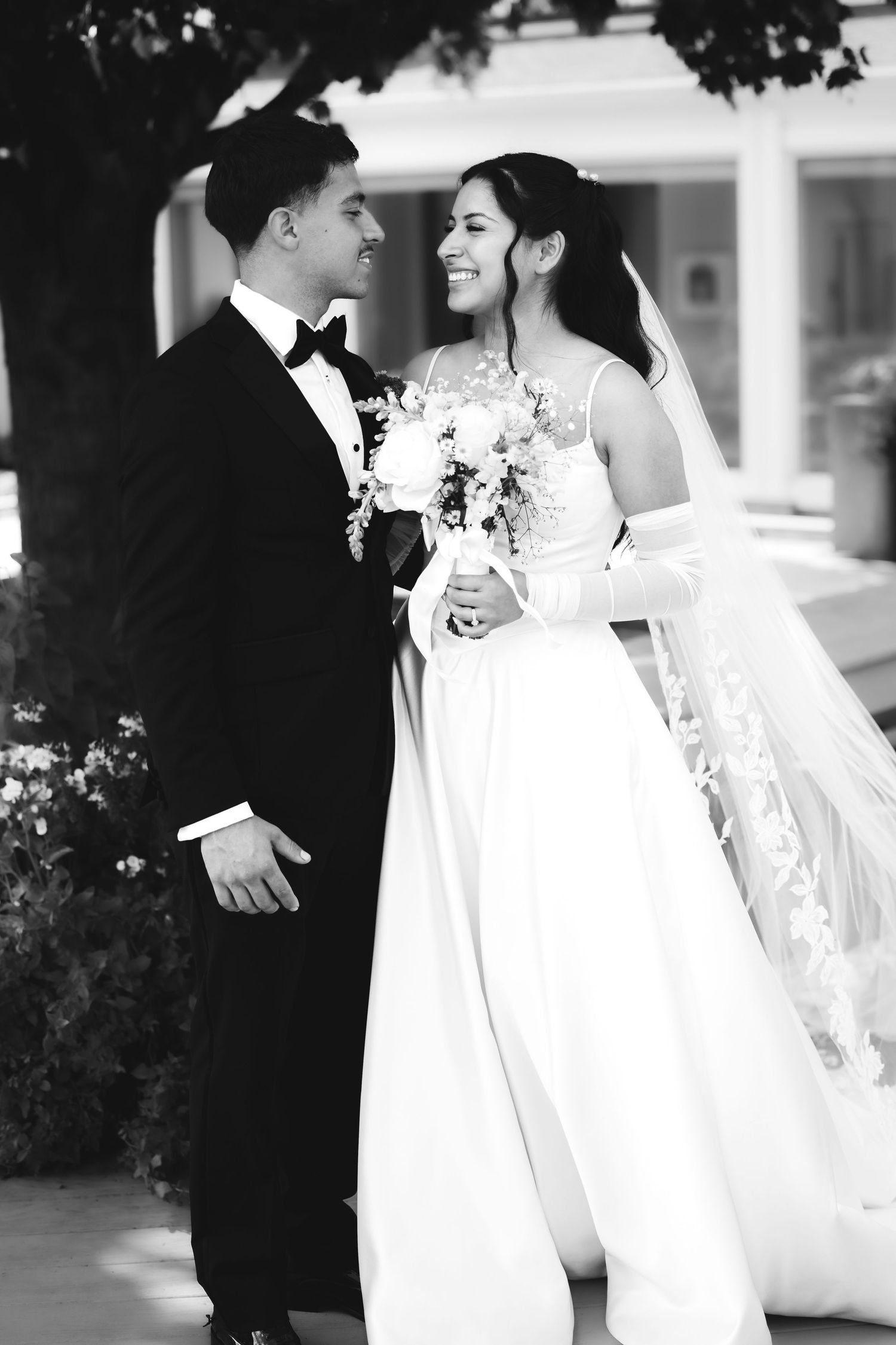 Black and white photo of smiling bride and groom in wedding attire standing together outdoors.