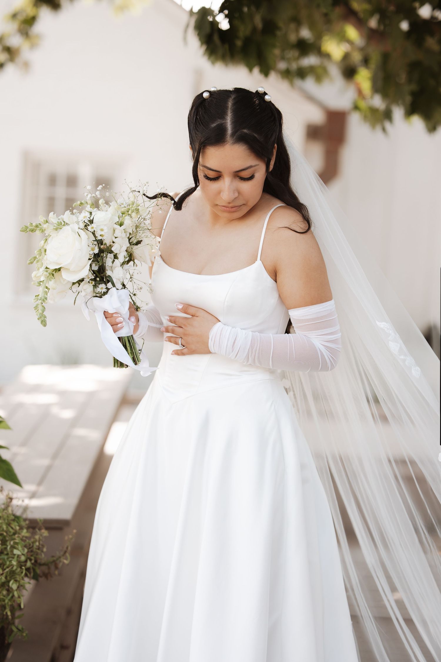Bride in white satin dress and veil holds white floral bouquet outdoors in sunlight.