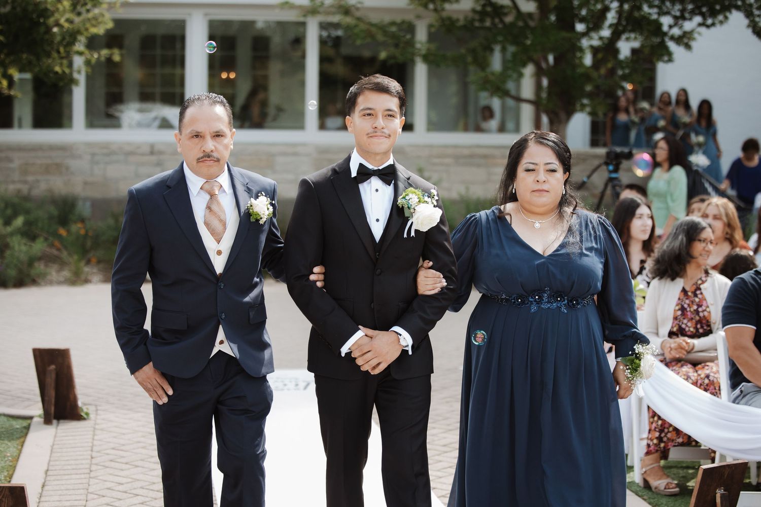 Groom in black tuxedo poses with parents in navy attire at outdoor wedding ceremony.