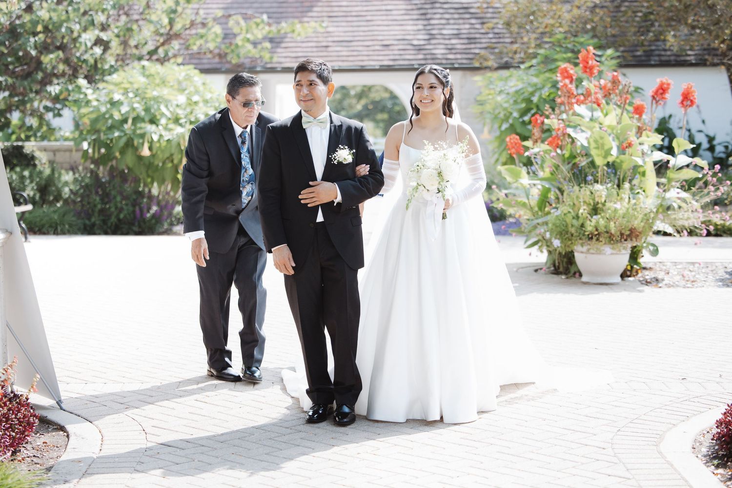 Bride in white ball gown is escorted down brick aisle by two men at outdoor ceremony.