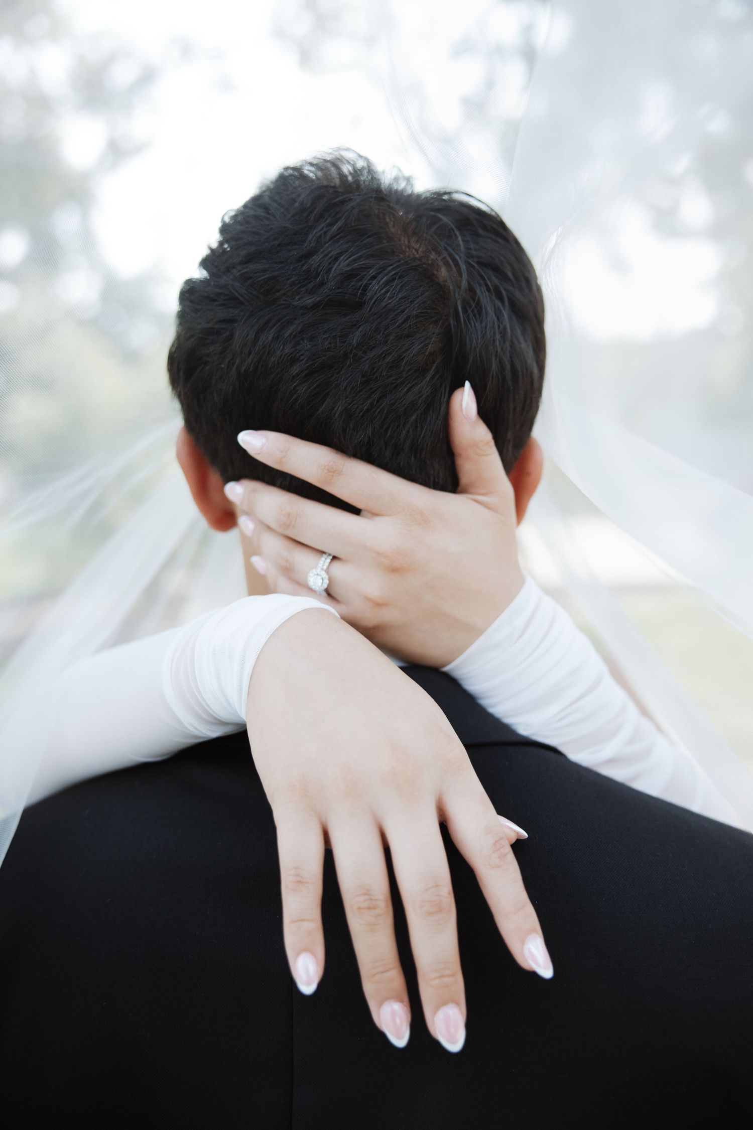 Bride's hand with diamond engagement ring resting on groom's shoulder during wedding embrace.
