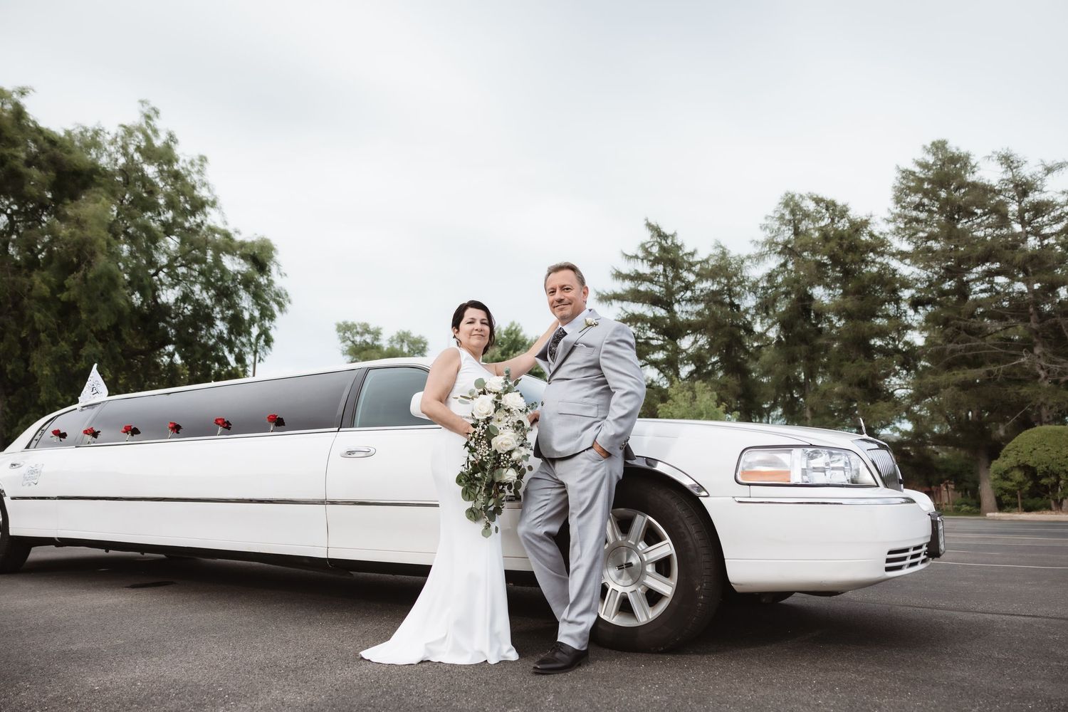 Bride and groom pose together in front of white stretch limousine on their wedding day outdoors.