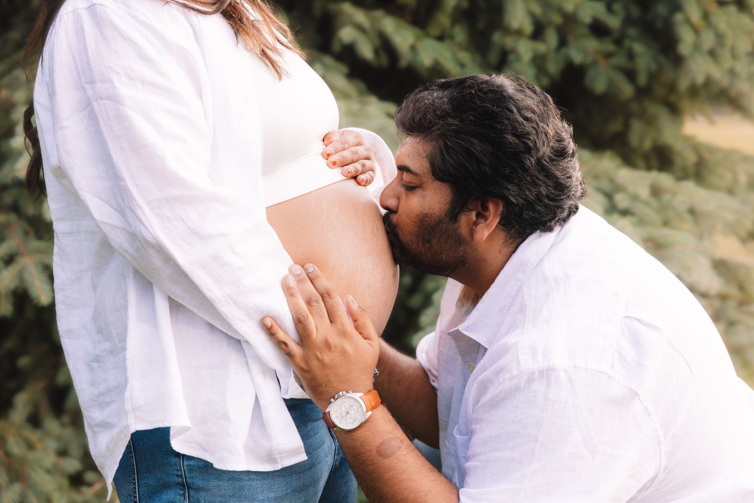 Man kneeling to kiss pregnant woman's bare baby bump outdoors near evergreen trees.