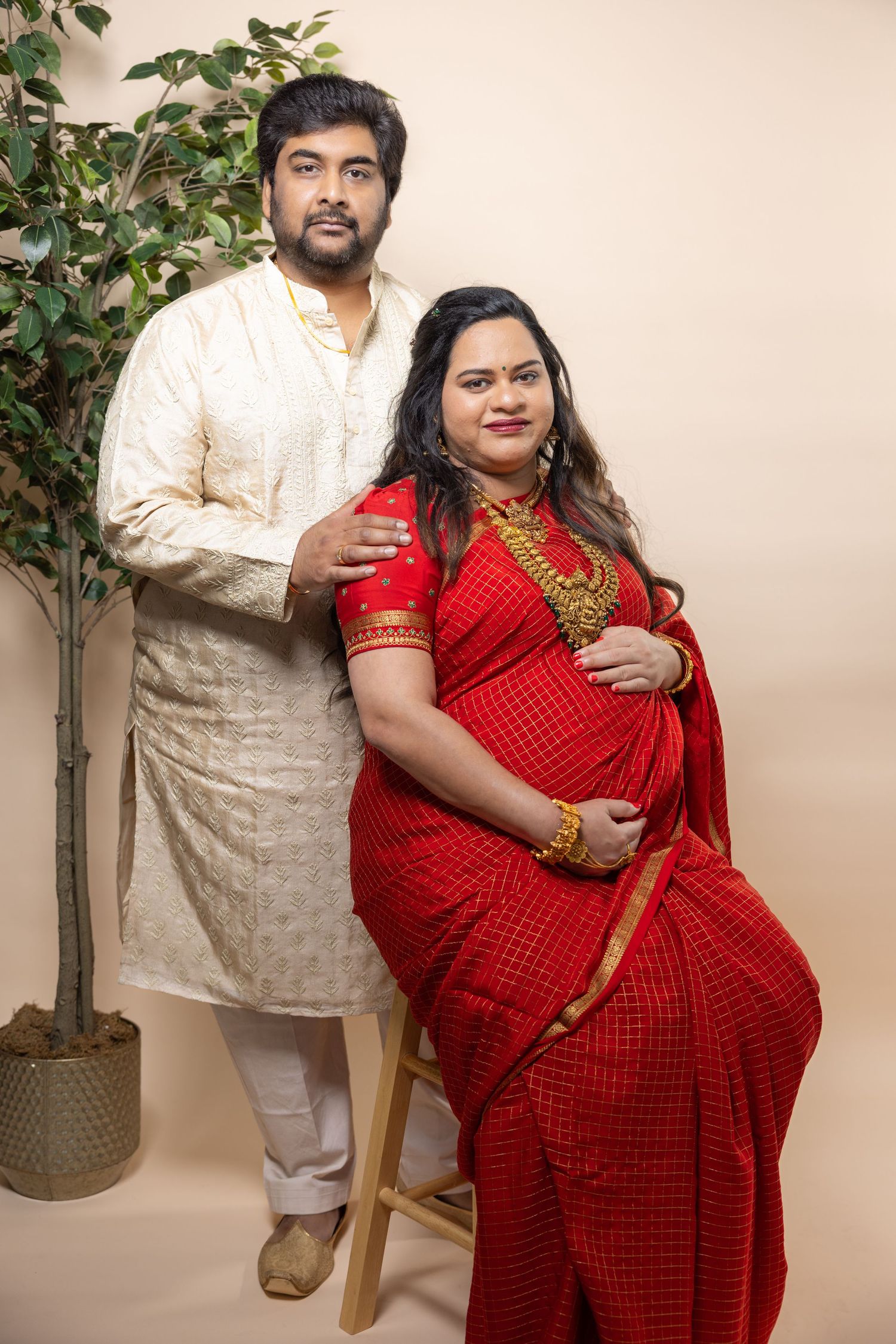Indian couple in traditional attire posing with pregnant woman in red saree seated on stool.