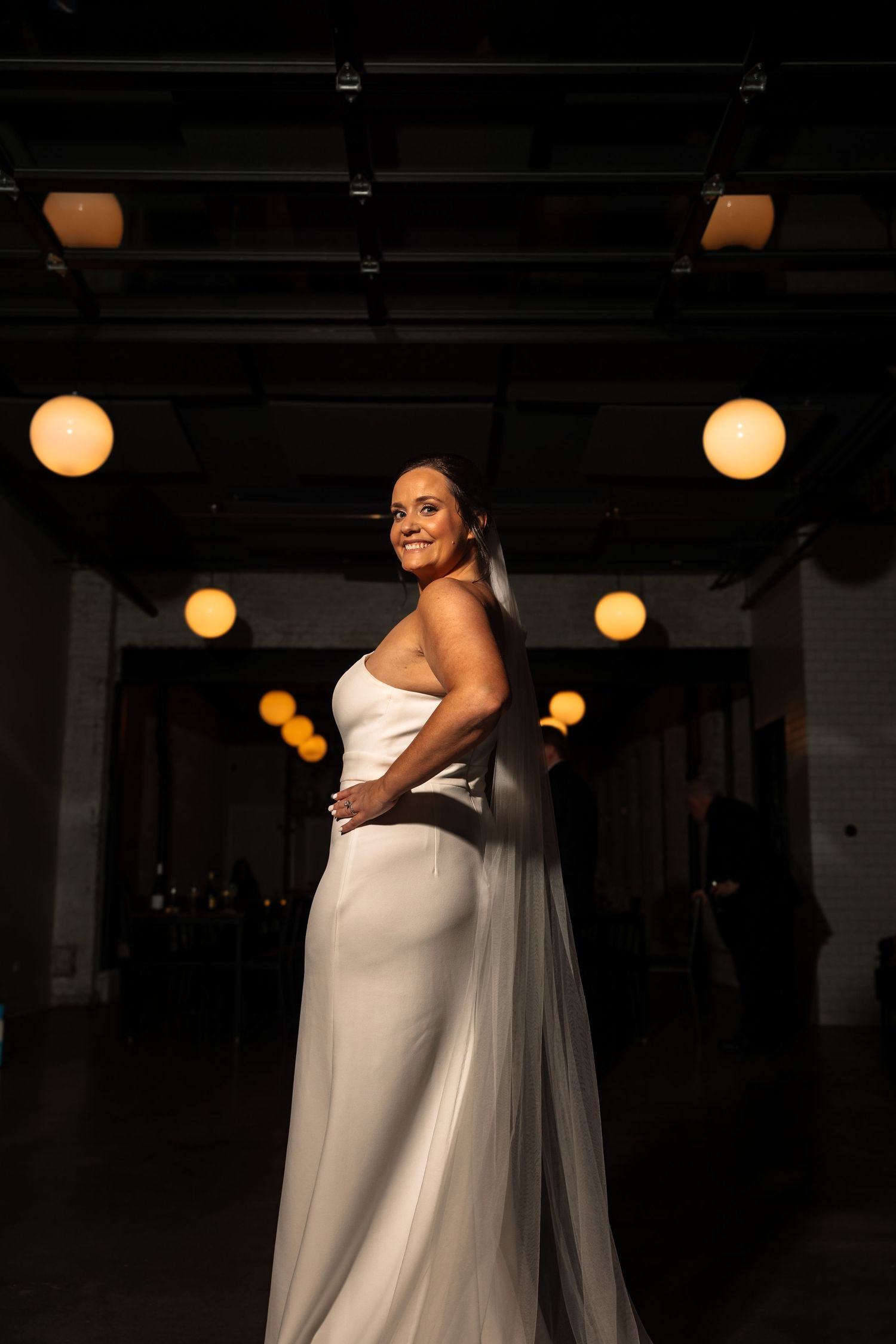 Elegant bride in minimalist white wedding dress smiles over shoulder in moody low-light industrial venue setting.