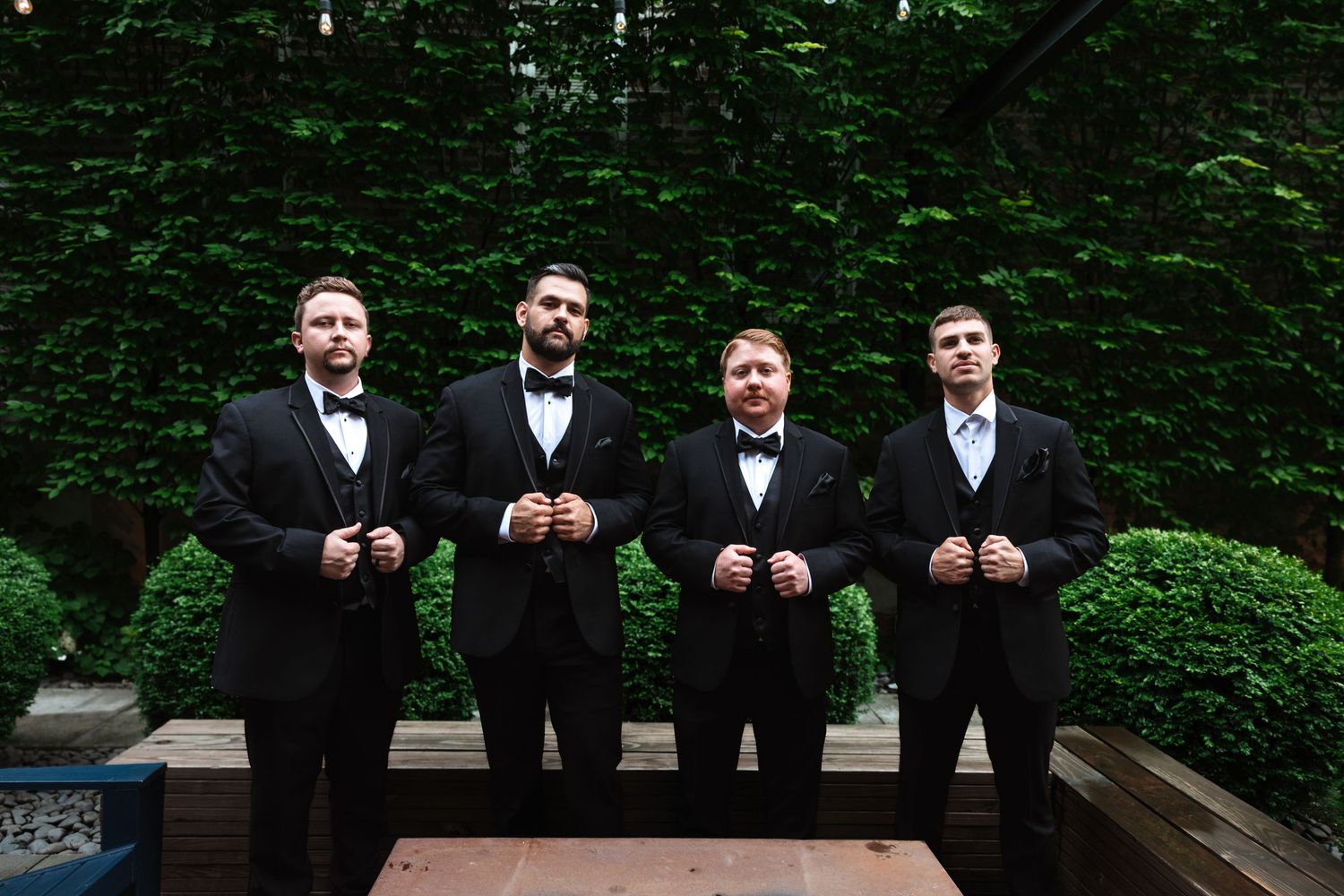 Four groomsmen in black tuxedos adjusting their lapels in front of a lush green hedge.