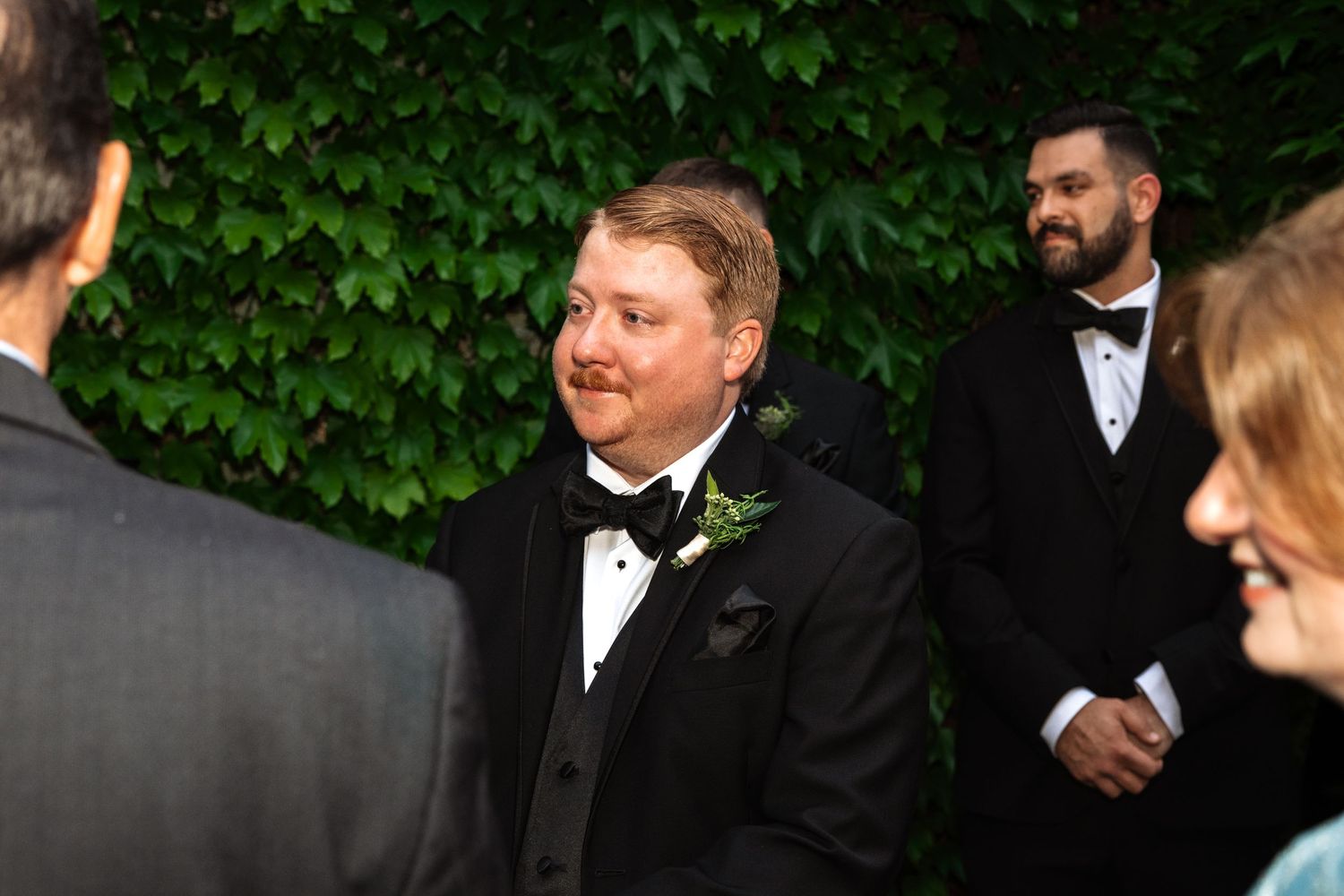 Groom in black tuxedo with boutonniere smiling at wedding ceremony outdoors with ivy wall backdrop.