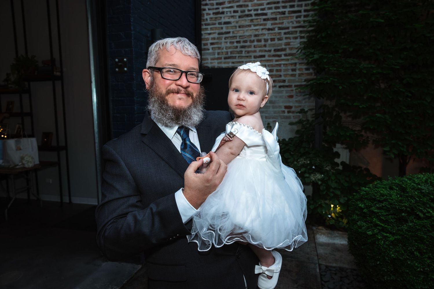 Bearded man in suit holding a baby girl in white dress at a wedding reception.