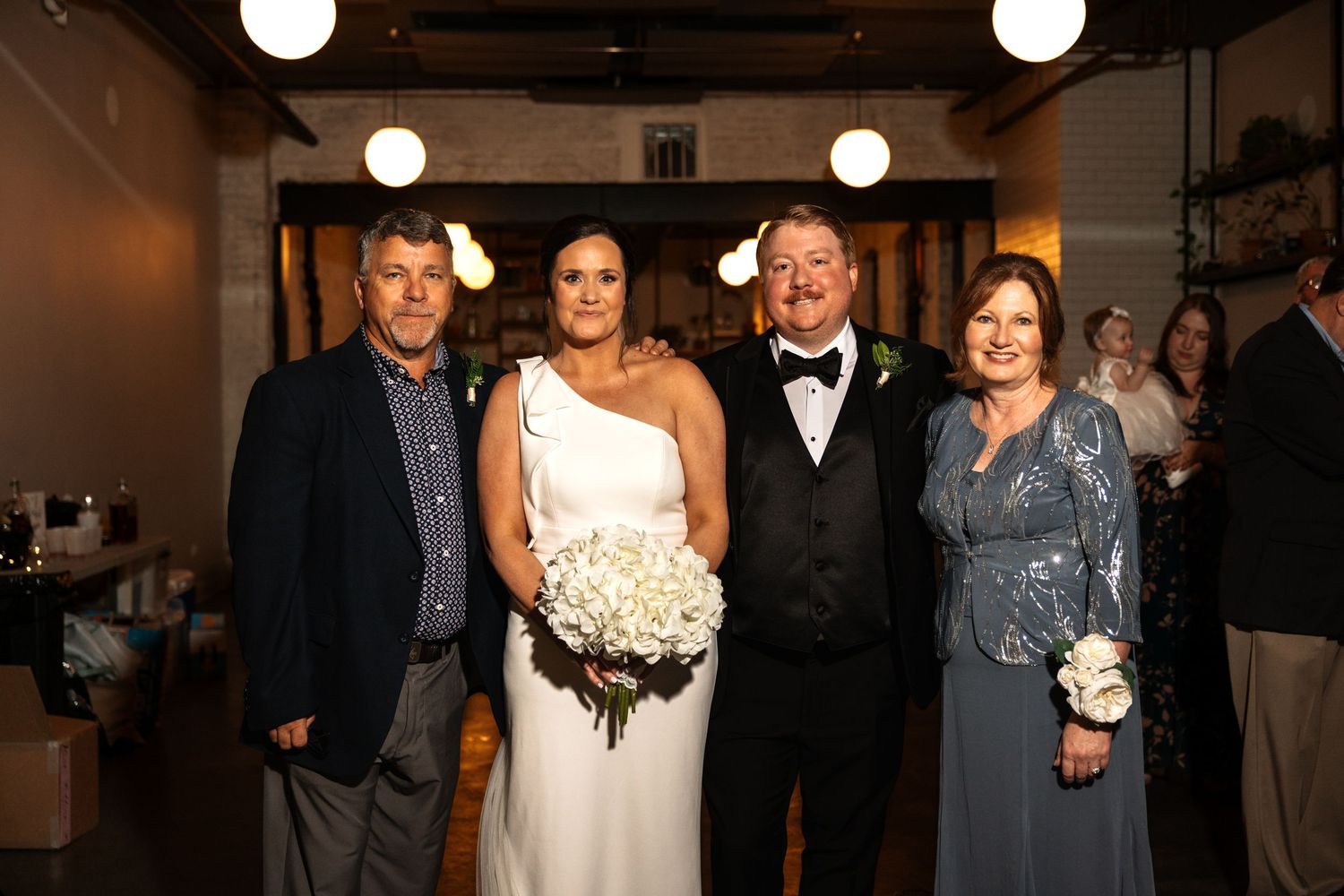 Bride holding white bouquet poses with groom in bow tie and two smiling family members at wedding reception.