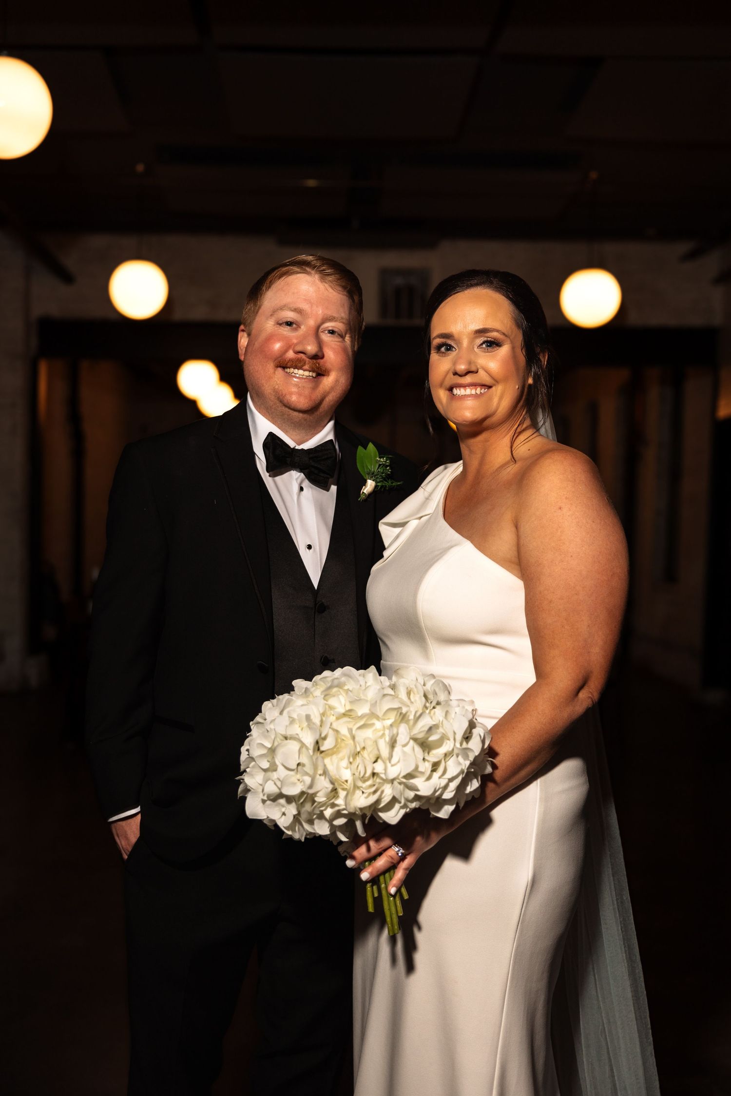 Bride in white gown and groom in tuxedo smile holding white hydrangea bouquet at wedding venue.