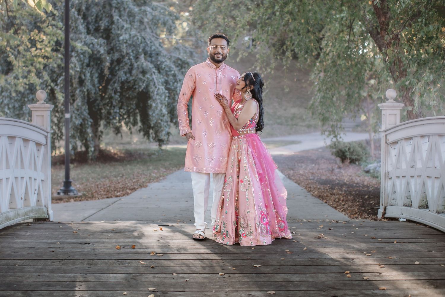 Indian couple in pink traditional attire stands romantically on white bridge in park.