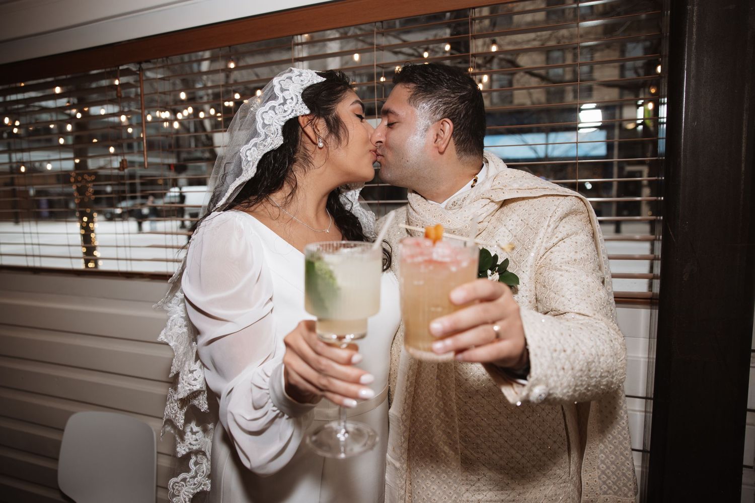Bride and groom kissing while holding cocktails at wedding reception, dressed in white bridal gown and ivory Indian sherwani.