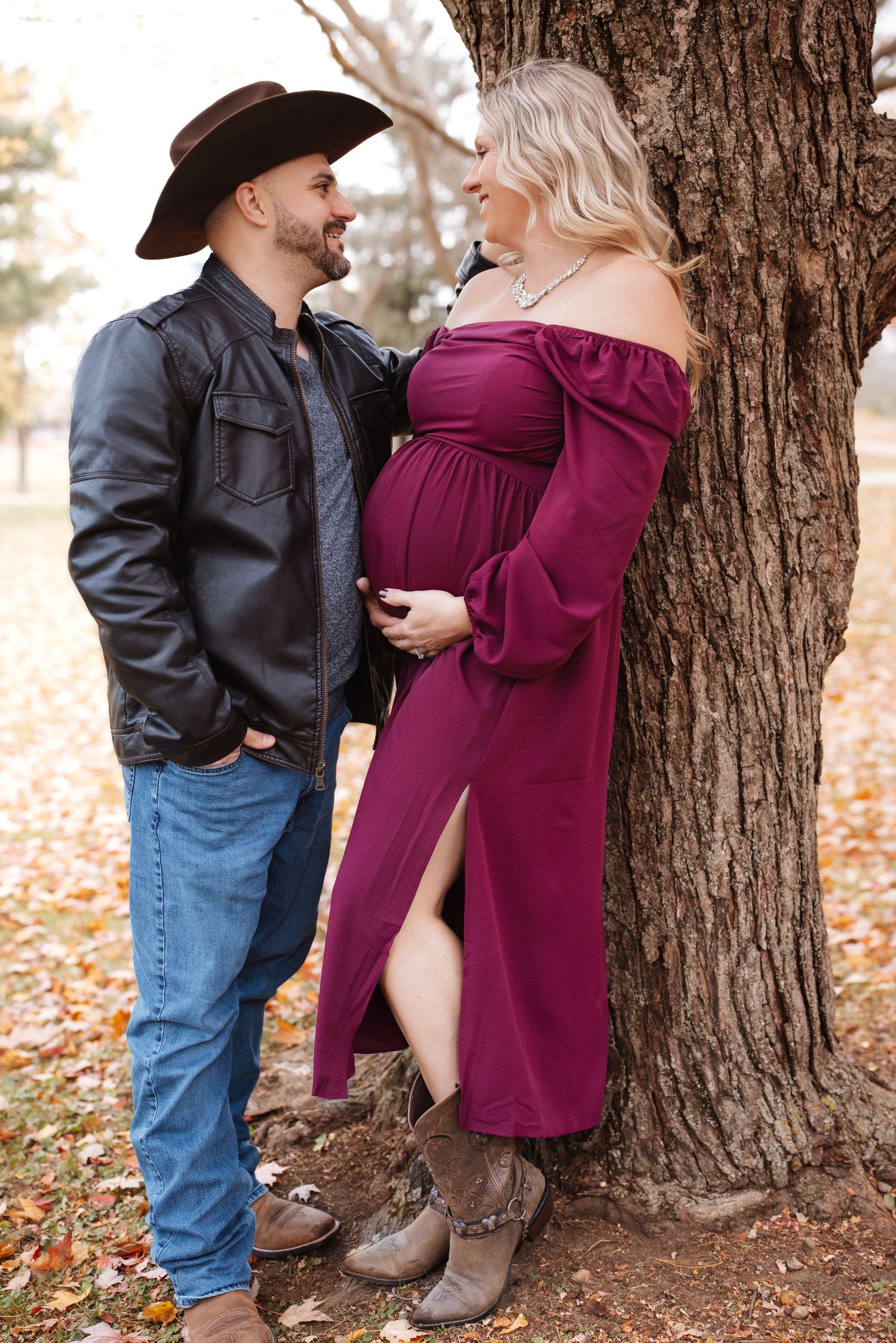 Couple sharing an intimate moment by a tree, man in cowboy hat with pregnant woman in burgundy maternity dress.