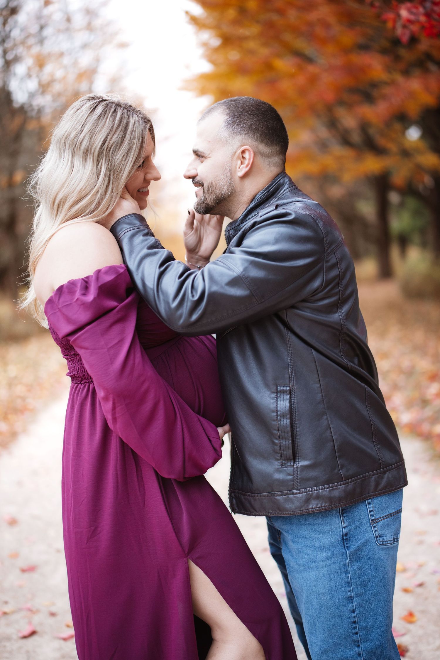 Couple sharing tender moment on fall foliage path, man in leather jacket holds womans chin as she smiles in purple maternity dress.