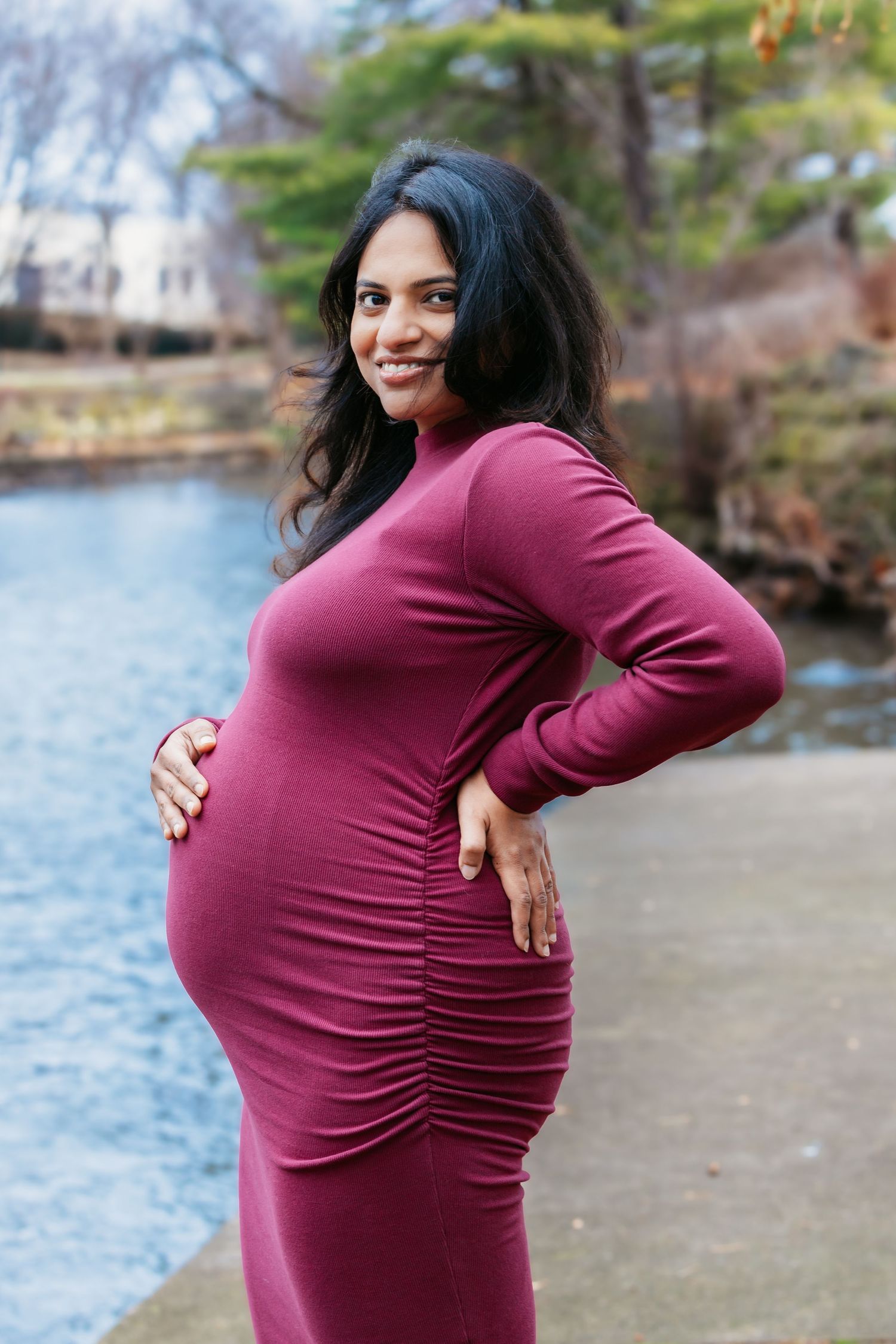 Smiling pregnant woman in burgundy ruched dress holding belly outdoors near water.