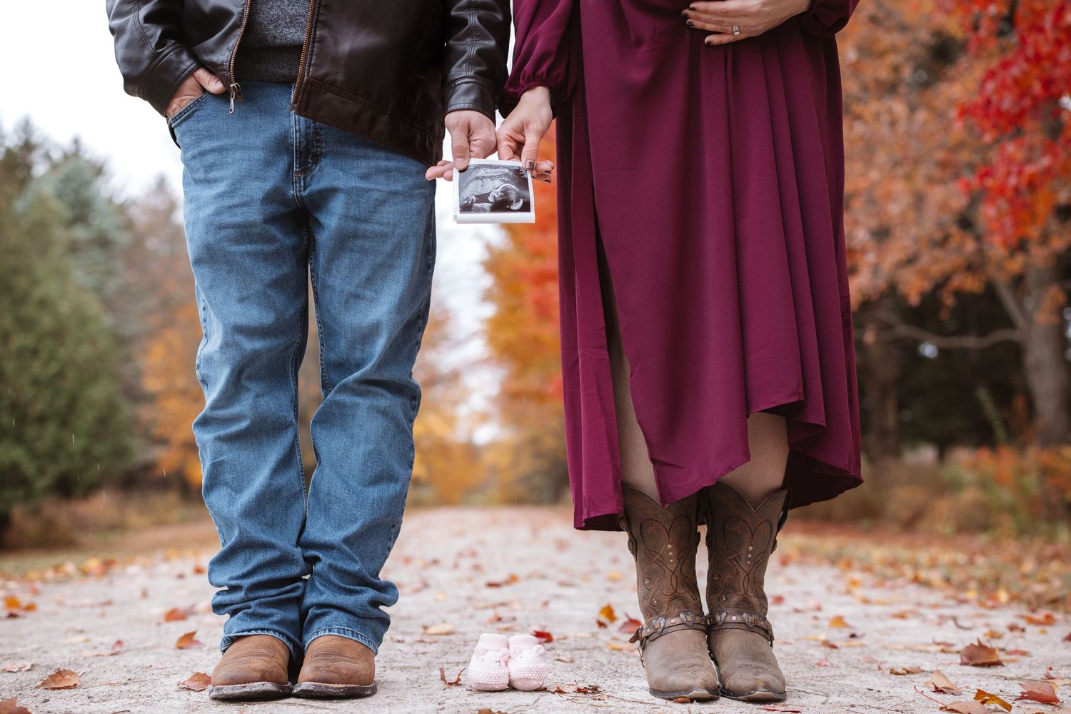 Couple holding ultrasound photo with tiny baby shoes on autumn path, pregnancy announcement.