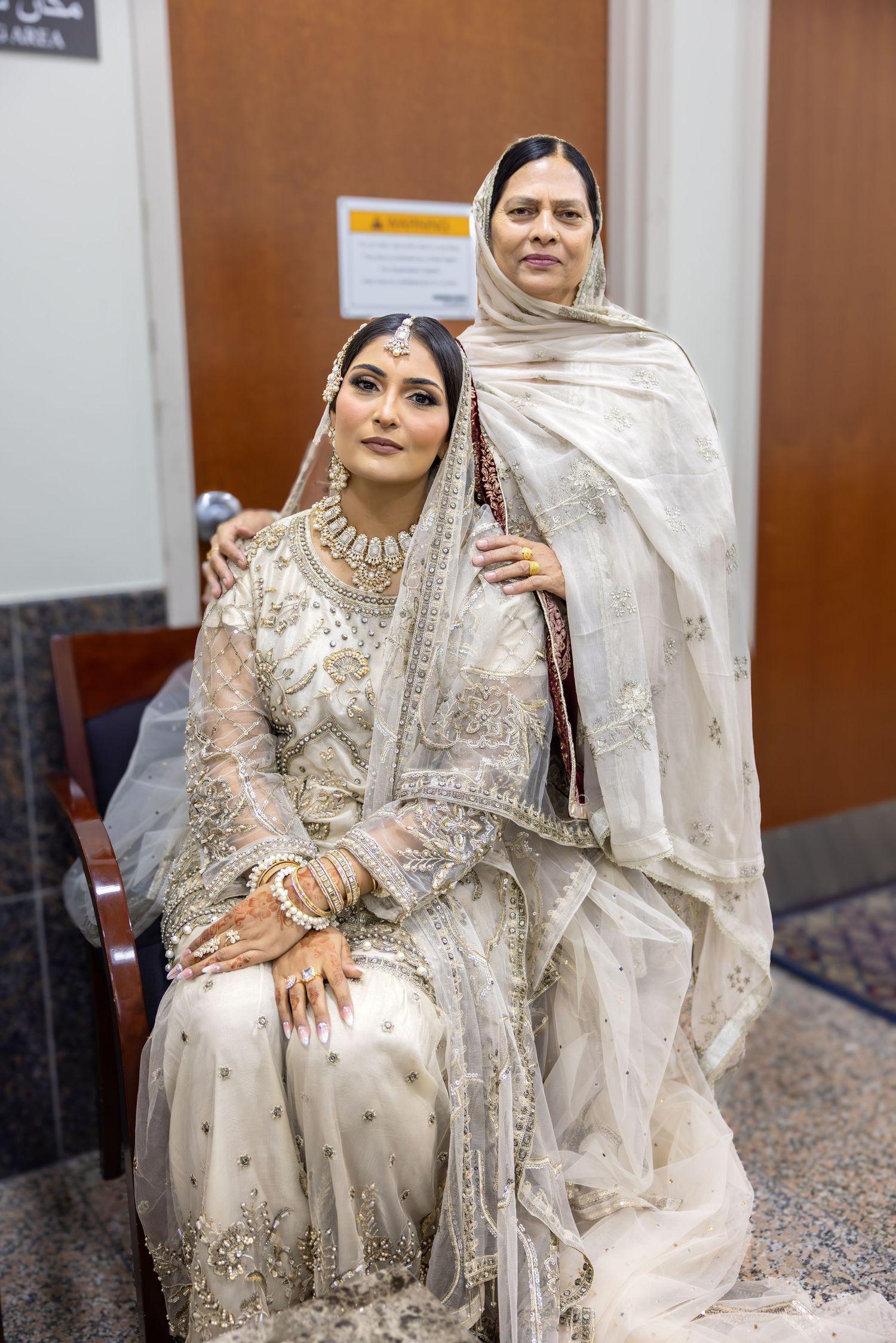 A seated South Asian bride in white embroidered bridal attire is embraced by an older woman in a white dupatta.